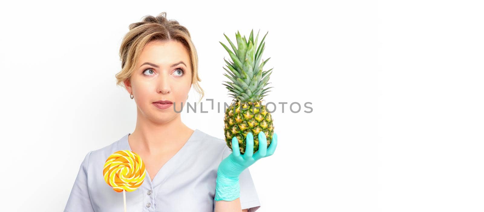 Young caucasian female doctor nutritionist holding fresh pineapple with lollipop over white background. by okskukuruza