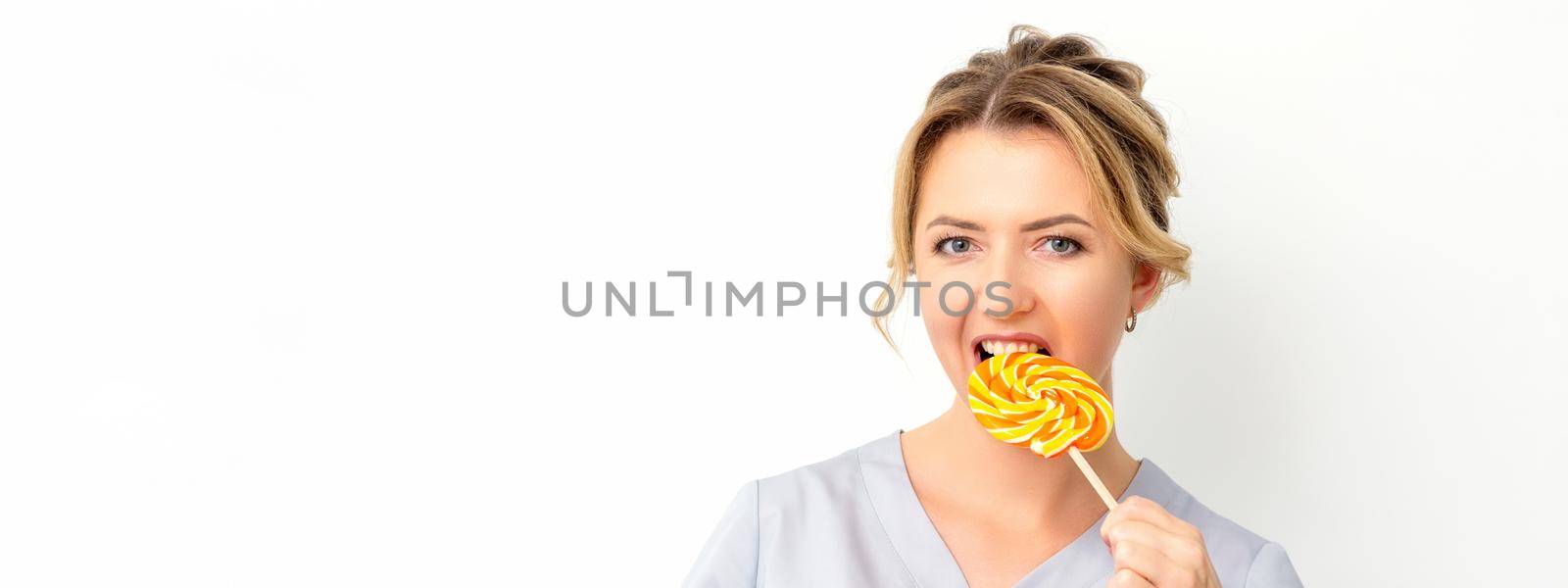 Portrait of a beautiful young caucasian beautician wearing a medical shirt bites a lollipop on a white background