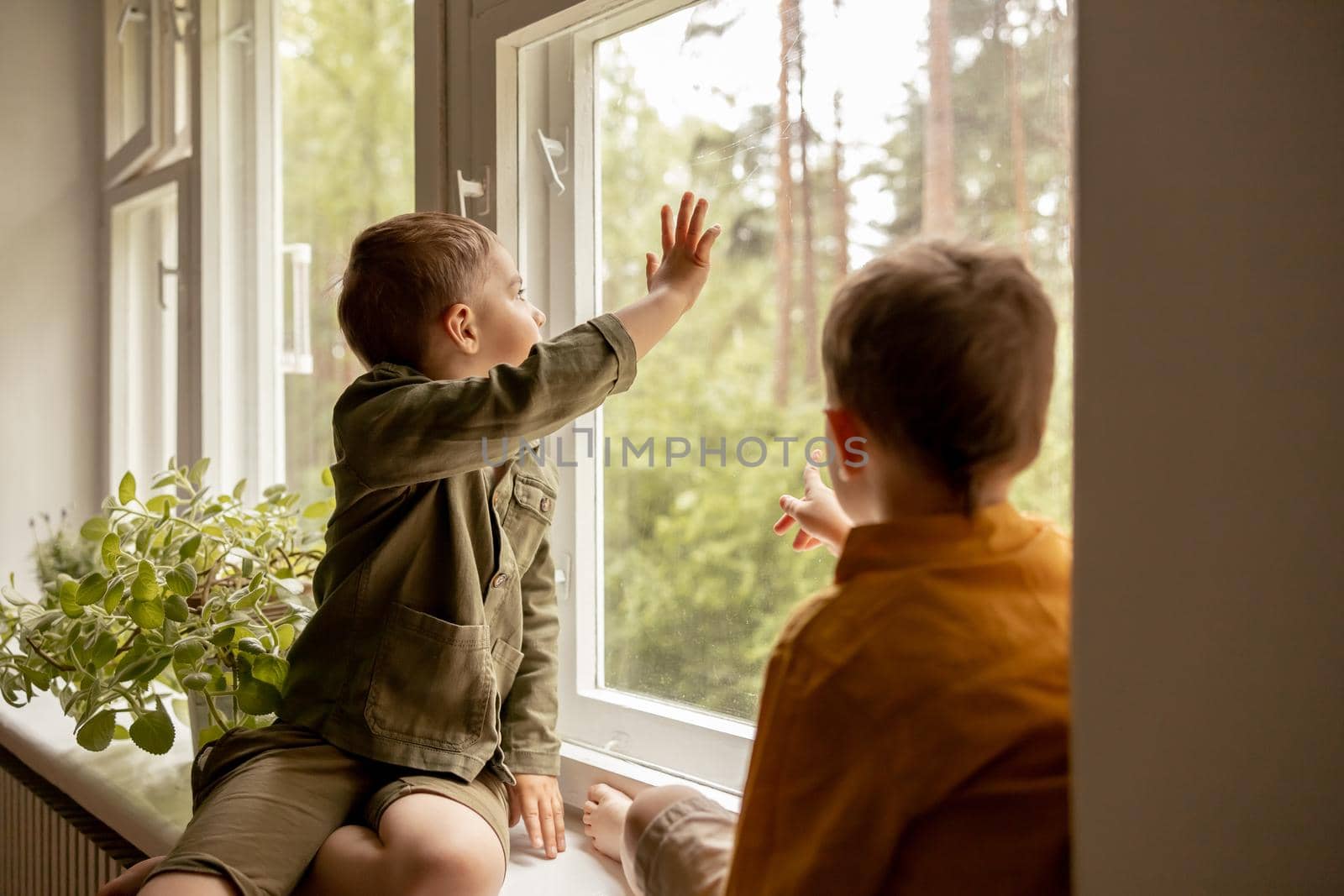 Children sitting on windowsill and waiting for someone comming. Two brothers, friends. Cute preschool kids alone at home. Boys are waiting for their mother or father. Loneliness. Busy parents
