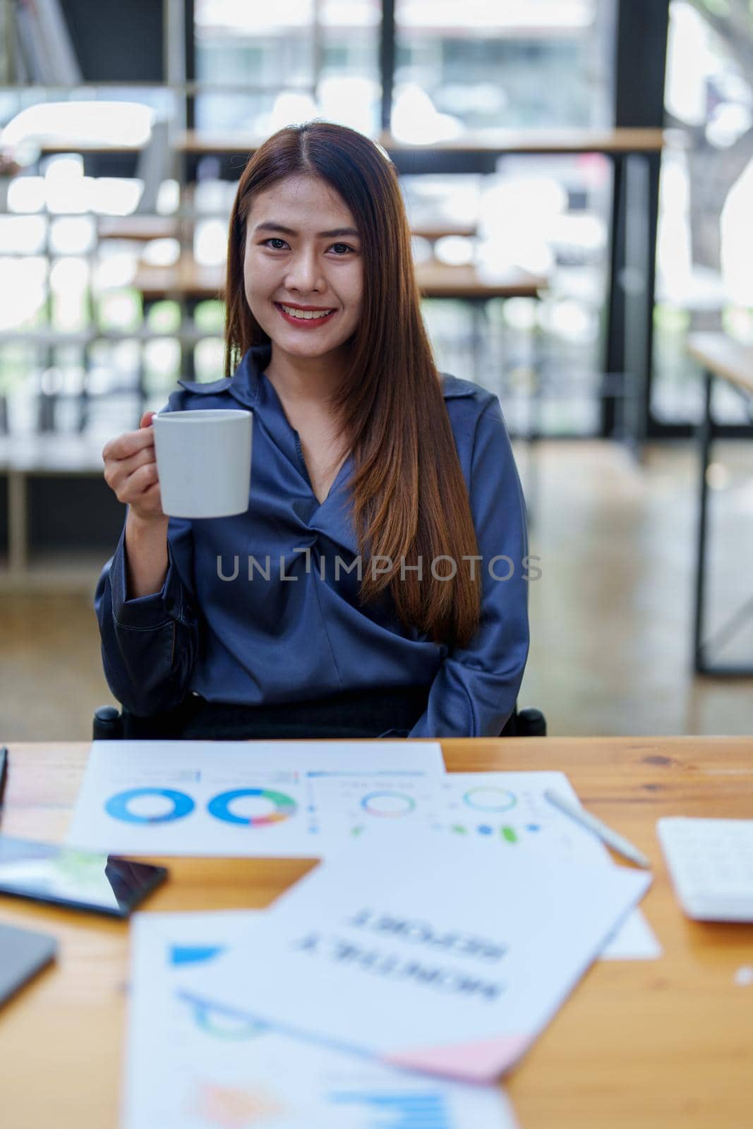 Portrait of a happy Asian woman smiling at her desk during the daytime coffee break by Manastrong