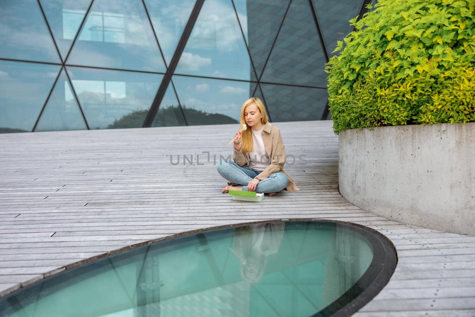 Young beautiful blond woman eating sushi outdoors, on the wooden terrace, by modern building in the city. Tasty food to go. Girl has lunch break, spending time outside, eating Asian food. City life