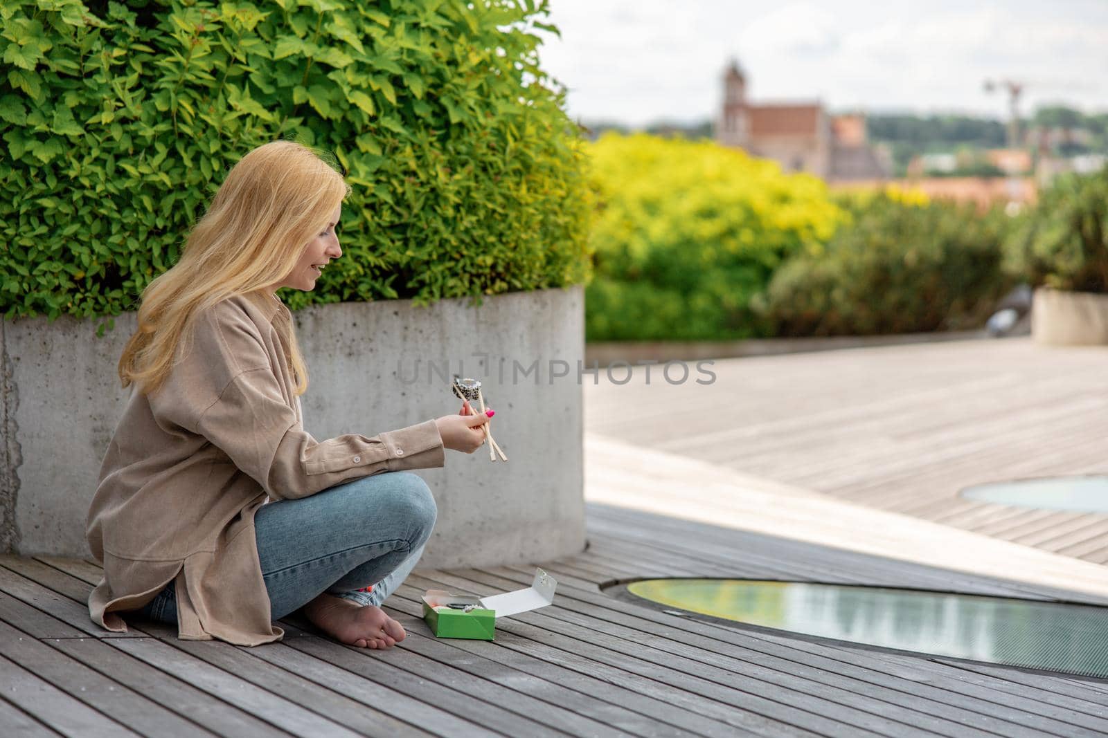 Young beautiful blond woman eating sushi outdoors, on the wooden terrace, by modern building in the city. Tasty food to go. Girl has lunch break, spending time outside, eating Asian food. City life
