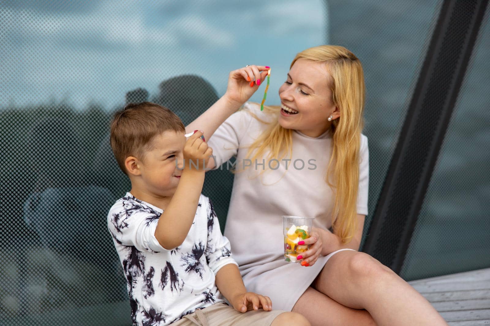 Happy blond woman and little boy sitting on terrace and eating sweets. Mother and son enjoy time together. Positive young mom playing, spending time with her cute child, laughing, having fun. Family