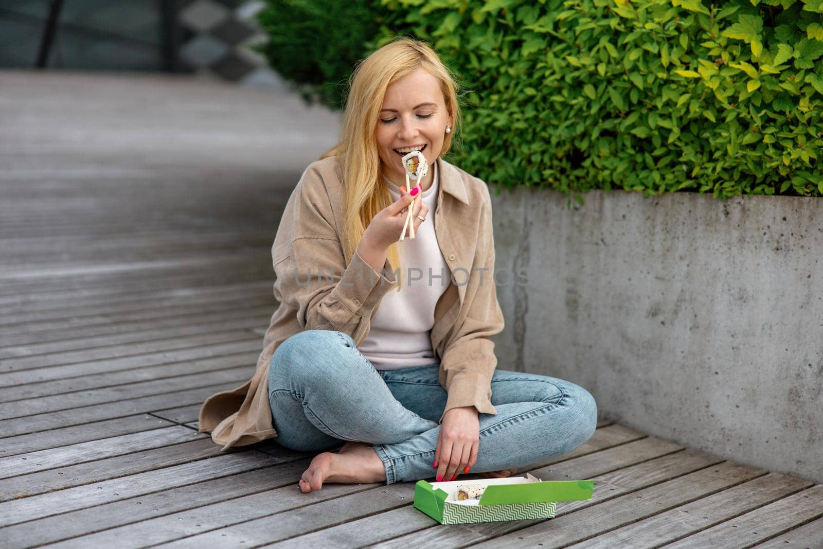 Young beautiful blond woman eating sushi outdoors, on the wooden terrace, by modern building in the city. Tasty food to go. Girl has lunch break, spending time outside and eating Asian food. City life by creativebird