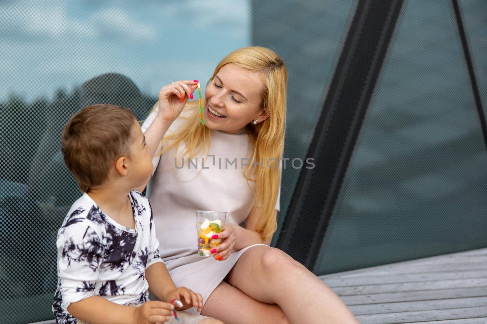 Happy blond woman and little boy sitting on terrace and eating sweets. Mother and son enjoy time together. Positive young mom playing, spending time with her cute child, laughing, having fun. Family. by creativebird
