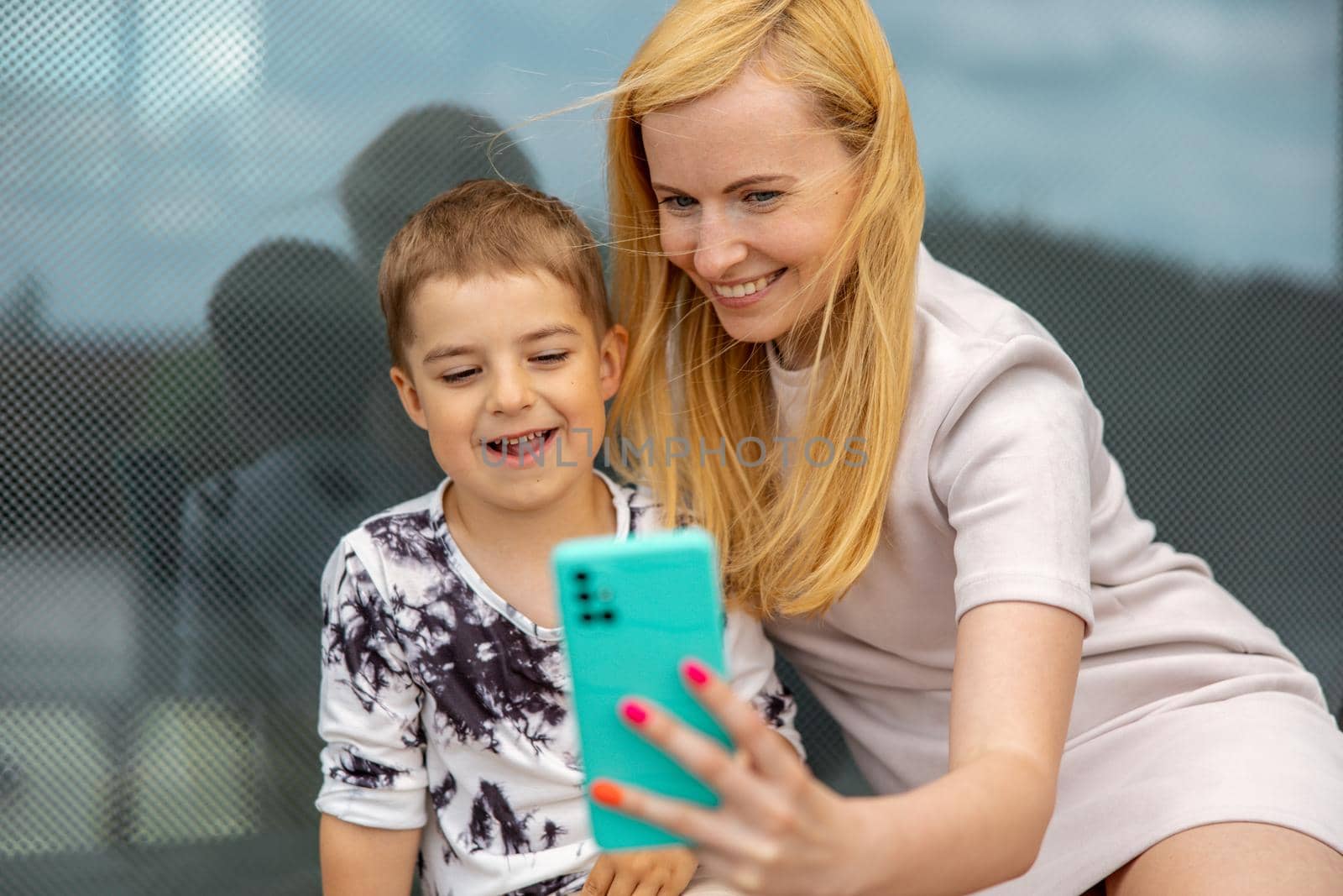 Happy blond woman and little boy sitting on terrace and making selfie on smartphone. Mother and son enjoy time together. Positive young mom spending time with her cute child. Having fun. Family. by creativebird