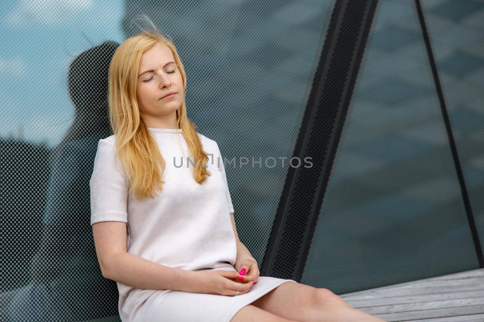 Young beautiful blond woman sitting outdoors, on the wooden terrace in the city and resting. Girl has break, spending time outside and relaxing. Time with yourself, dreaming, relaxation, mental health by creativebird
