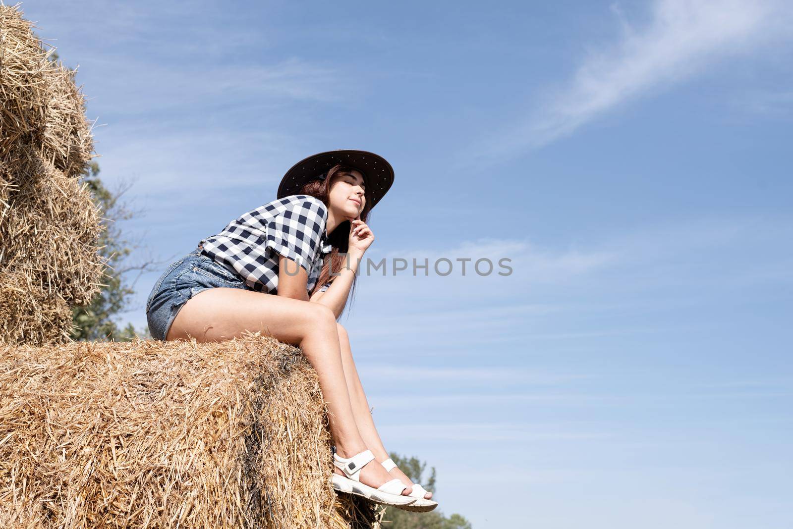 beautiful woman in plaid shirt and cowboy hat resting on haystack against sky background by Desperada