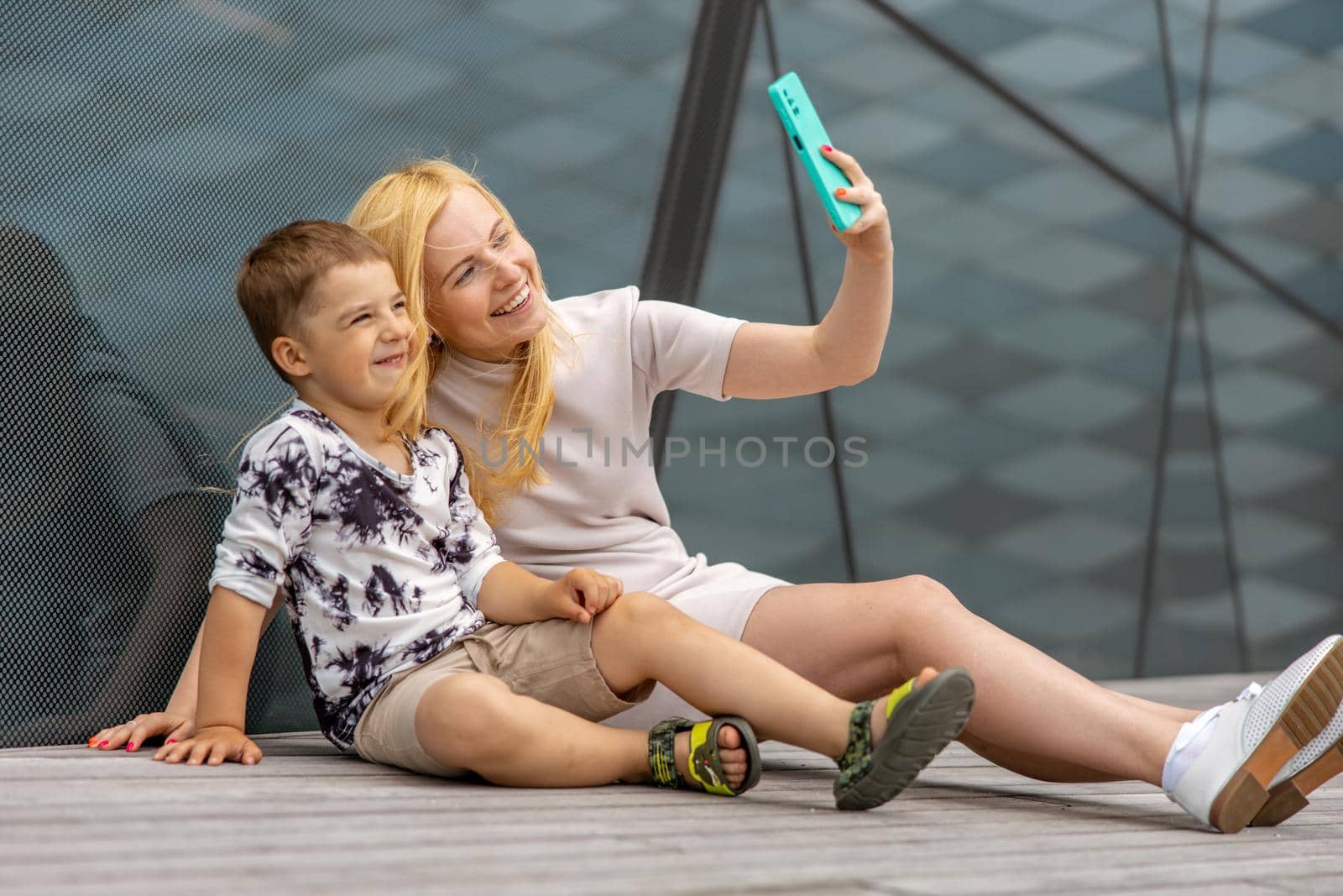Happy blond woman and little boy sitting on terrace and making selfie on smartphone. Mother and son enjoy time together. Positive young mom spending time with her cute child. Having fun. Family. by creativebird
