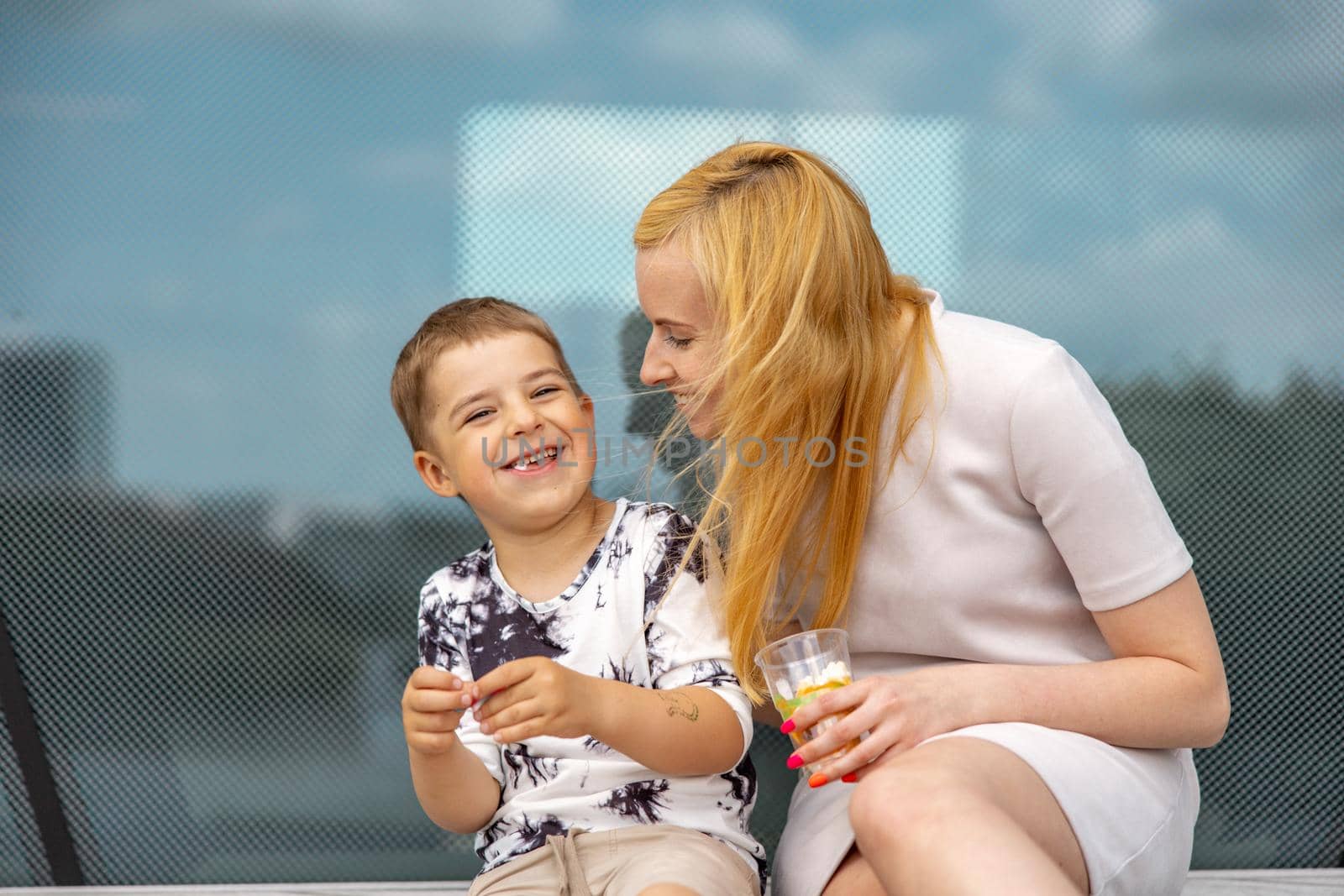 Happy blond woman and little boy sitting on terrace and eating sweets. Mother and son enjoy time together. Positive young mom playing, spending time with her cute child, laughing, having fun. Family. by creativebird