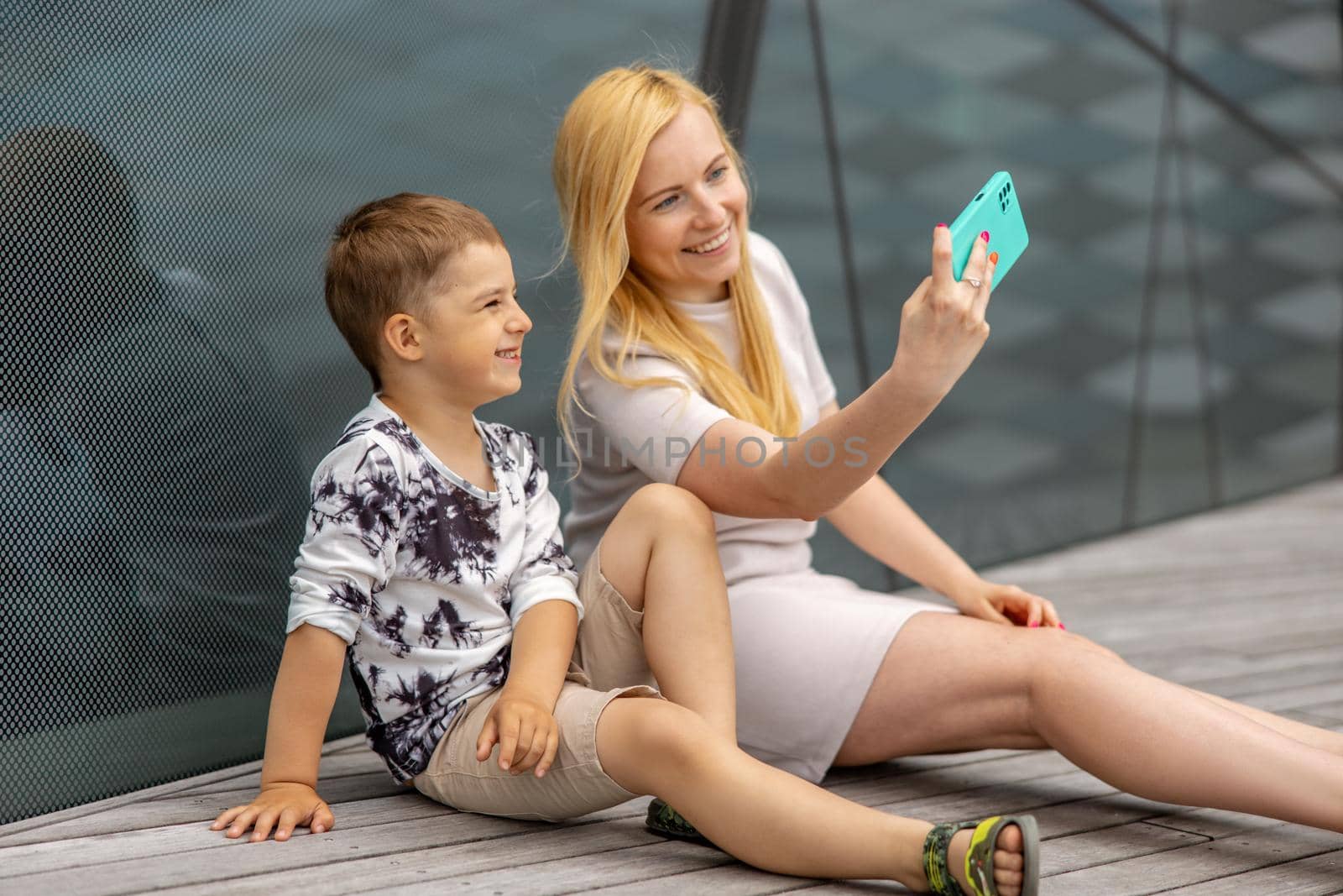 Happy blond woman and little boy sitting on terrace and making selfie on smartphone. Mother and son enjoy time together. Positive young mom spending time with her cute child. Having fun. Family