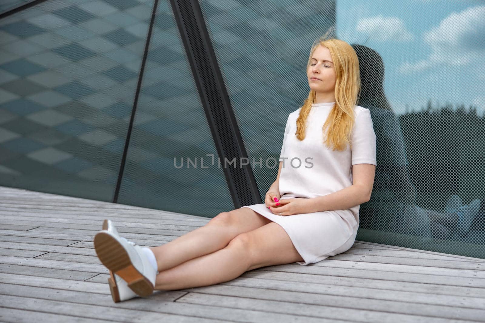 Young beautiful blond woman sitting outdoors, on the wooden terrace in the city and resting. Girl has break, spending time outside, relaxing. Time with yourself, dreaming, relaxation, mental health