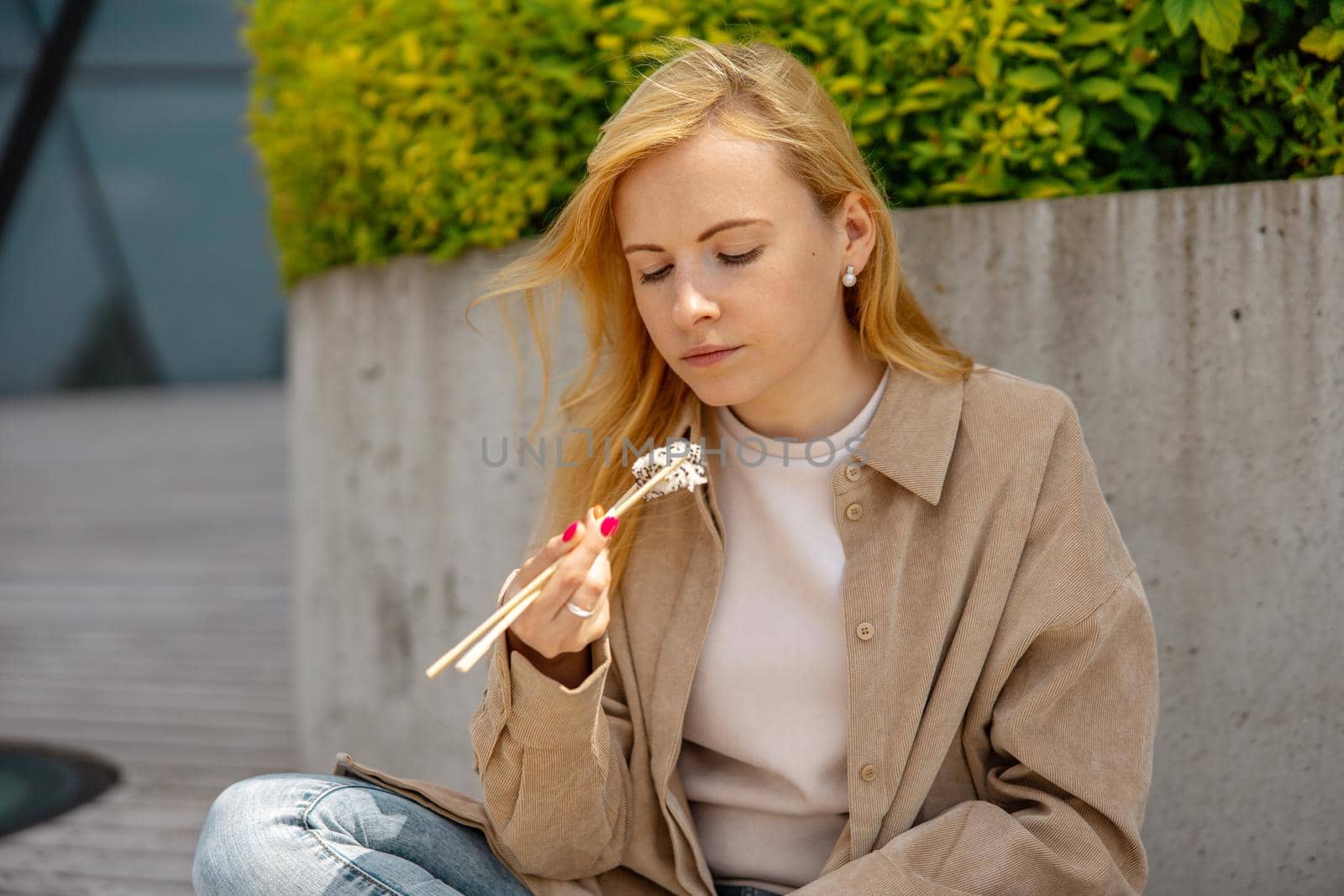 Young beautiful blond woman eating sushi outdoors, on the wooden terrace, by modern building in the city. Tasty food to go. Girl has lunch break, spending time outside and eating Asian food. City life by creativebird