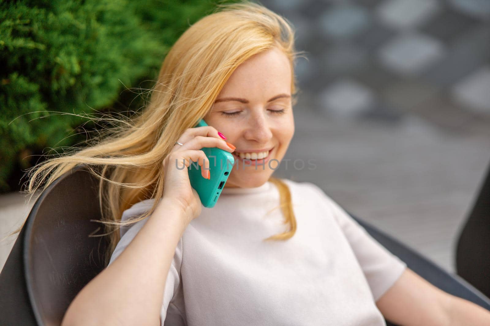 Young blond woman sittings outdoors in the city and making a call with her smartphone. Girl talking with friends with mobile phone. Leisure activity, communication. Conversation. Mobile technology