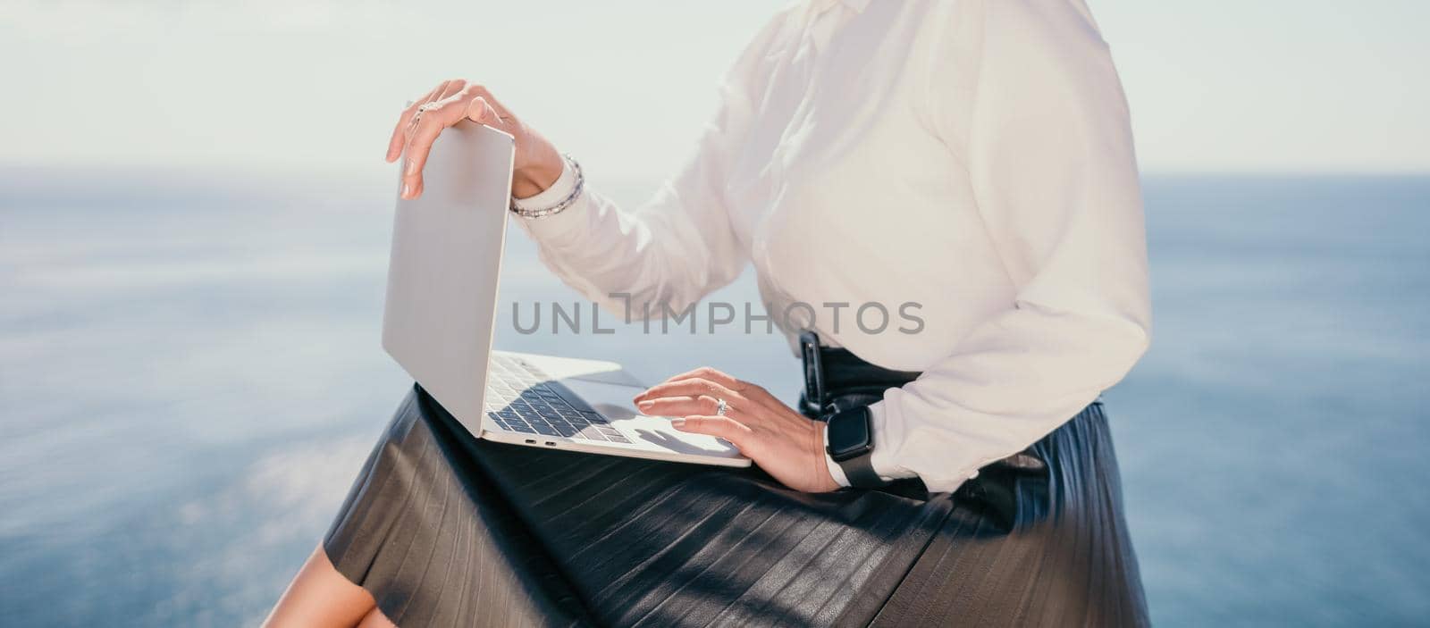 Successful business woman in yellow hat working on laptop by the sea. Pretty lady typing on computer at summer day outdoors. Freelance, travel and holidays concept.