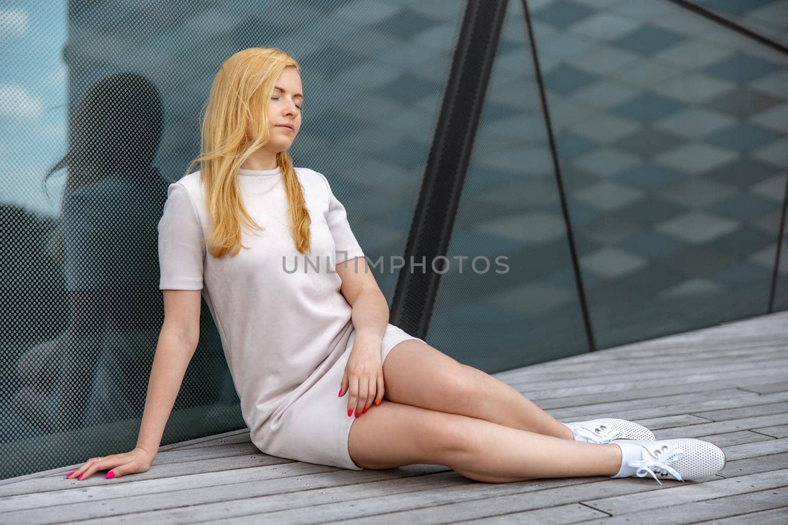 Young beautiful blond woman sitting outdoors, on the wooden terrace in the city and resting. Girl has break, spending time outside and relaxing. Time with yourself, dreaming, relaxation, mental health by creativebird