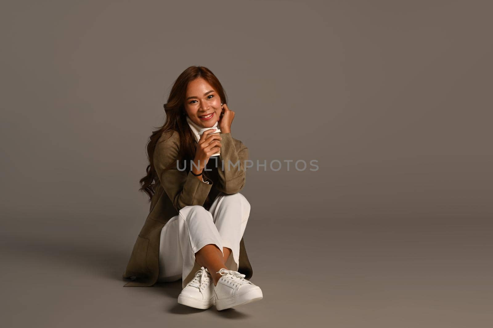 Portrait of stylish young woman in trendy coat holding paper cup sitting over grey background. Autumn and Winter concept.