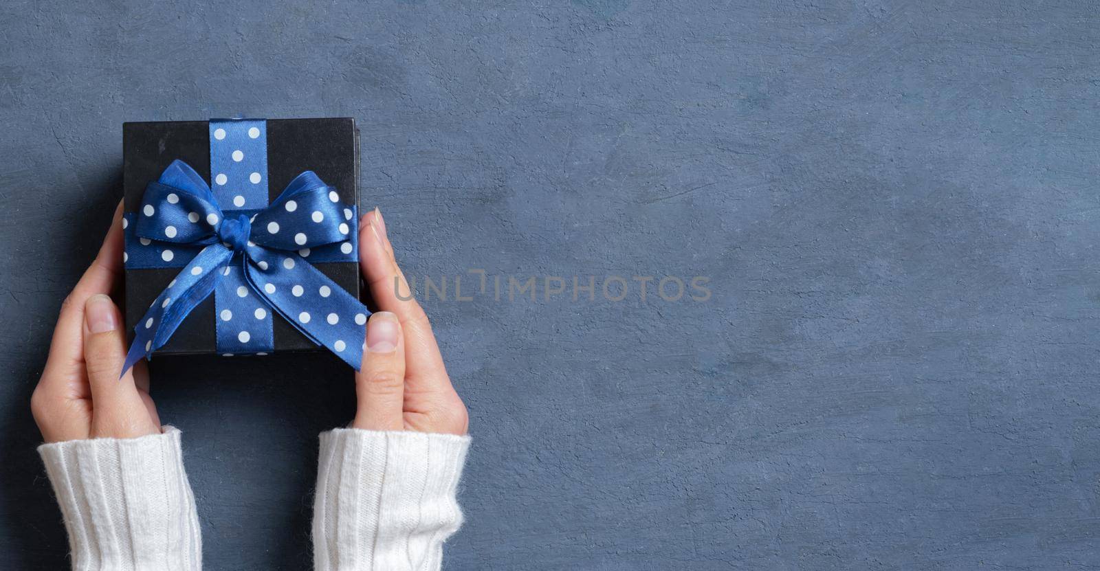 Black gift box with bow in female hands flat lay on dark cement background. Top view. Copy space.