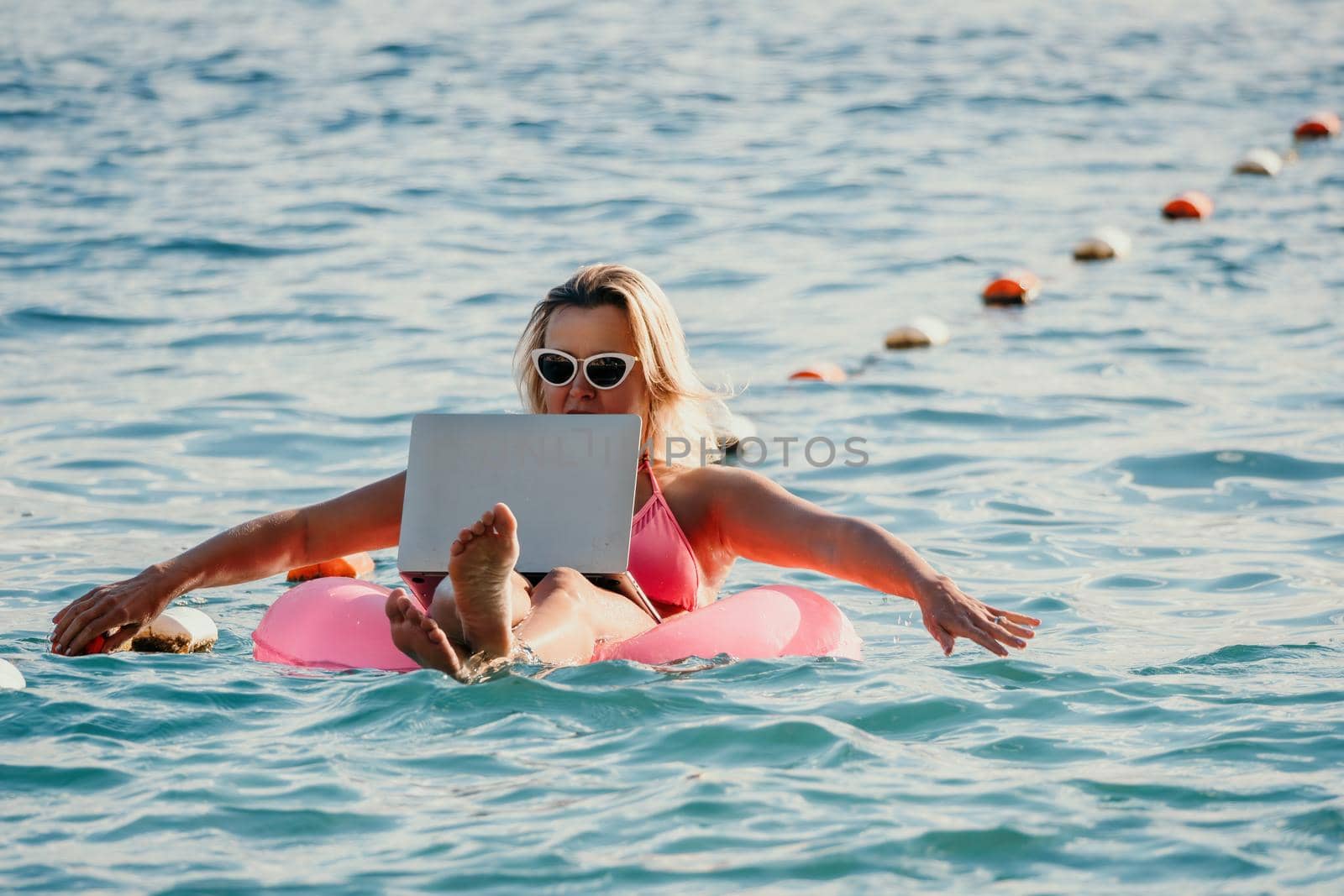 Freelancer, young blond woman in sunglases floating on an inflatable big pink donut with a laptop in the sea at sunset. Freelance, travel and holidays concept. Slow motion