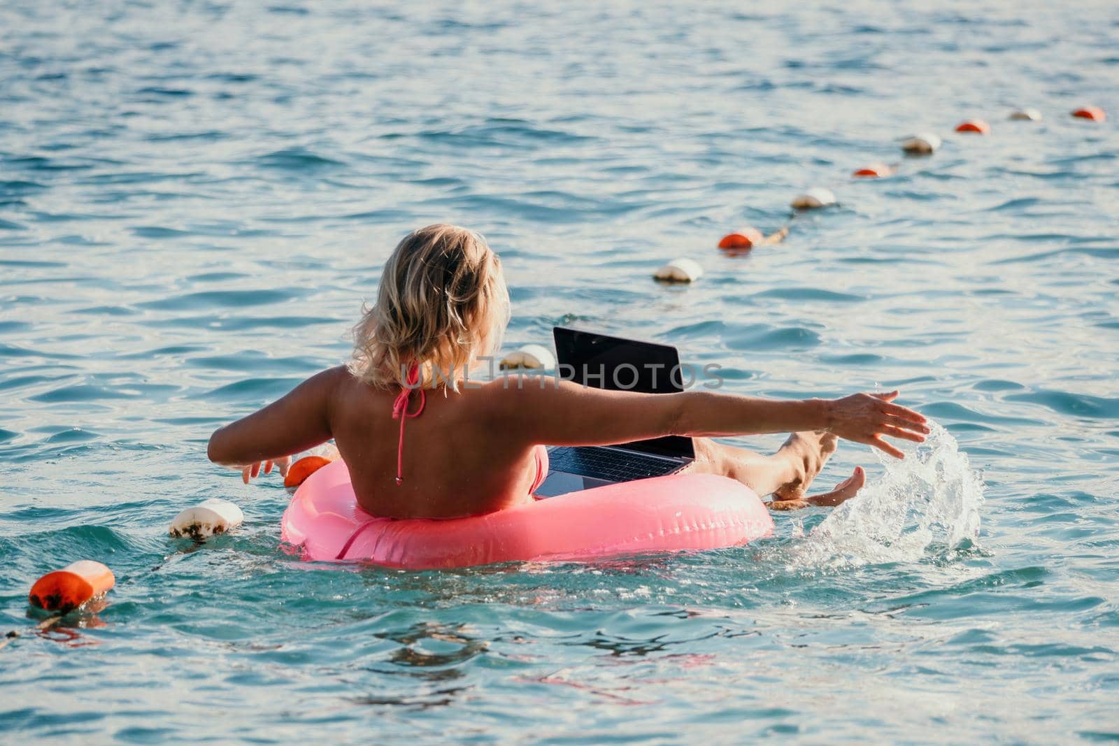Freelancer, young blond woman in sunglases floating on an inflatable big pink donut with a laptop in the sea at sunset. Freelance, travel and holidays concept. Slow motion