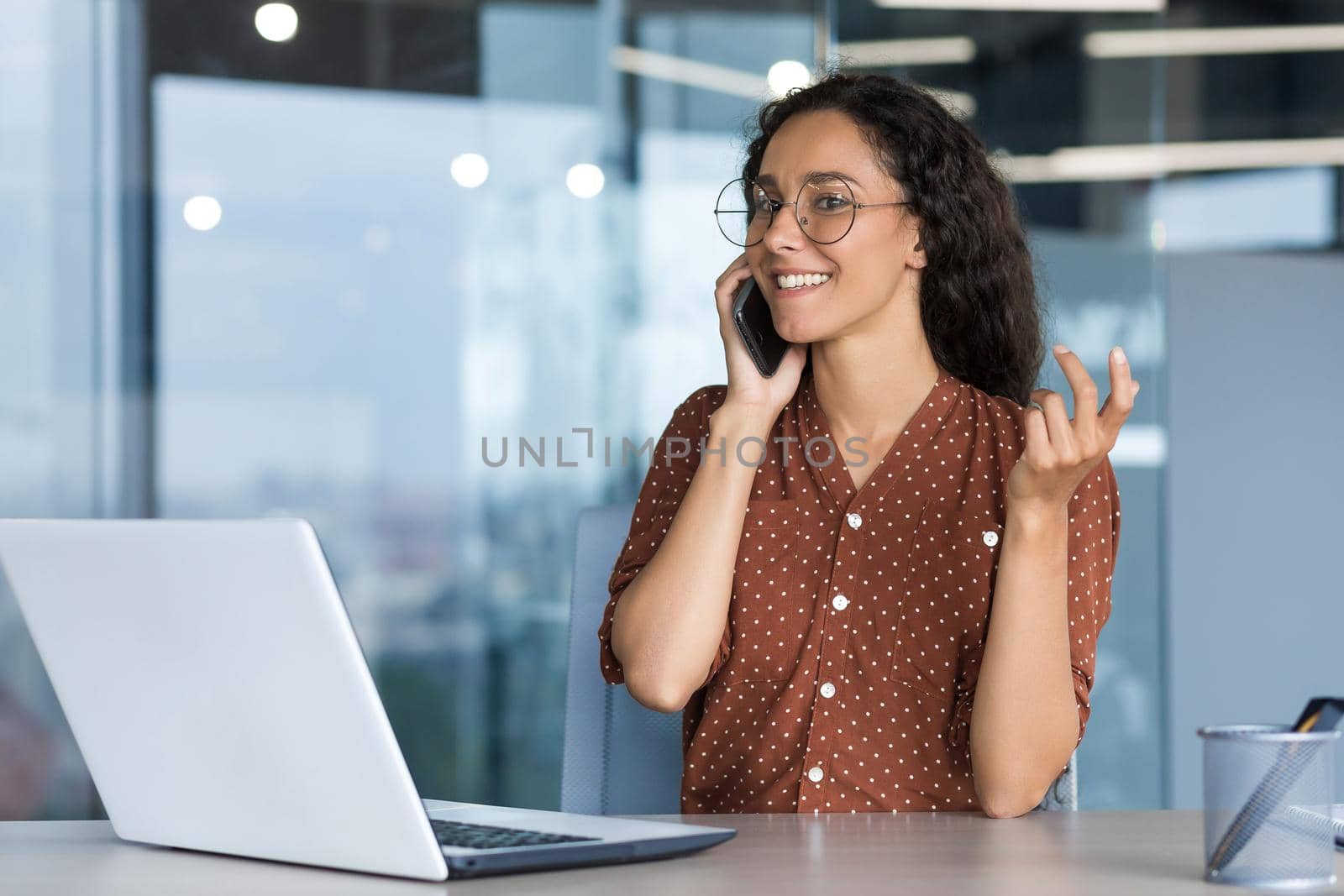 Young successful hispanic businesswoman working inside modern office, female employee cheerfully talking to colleagues on phone and smiling, using laptop while sitting at desk.