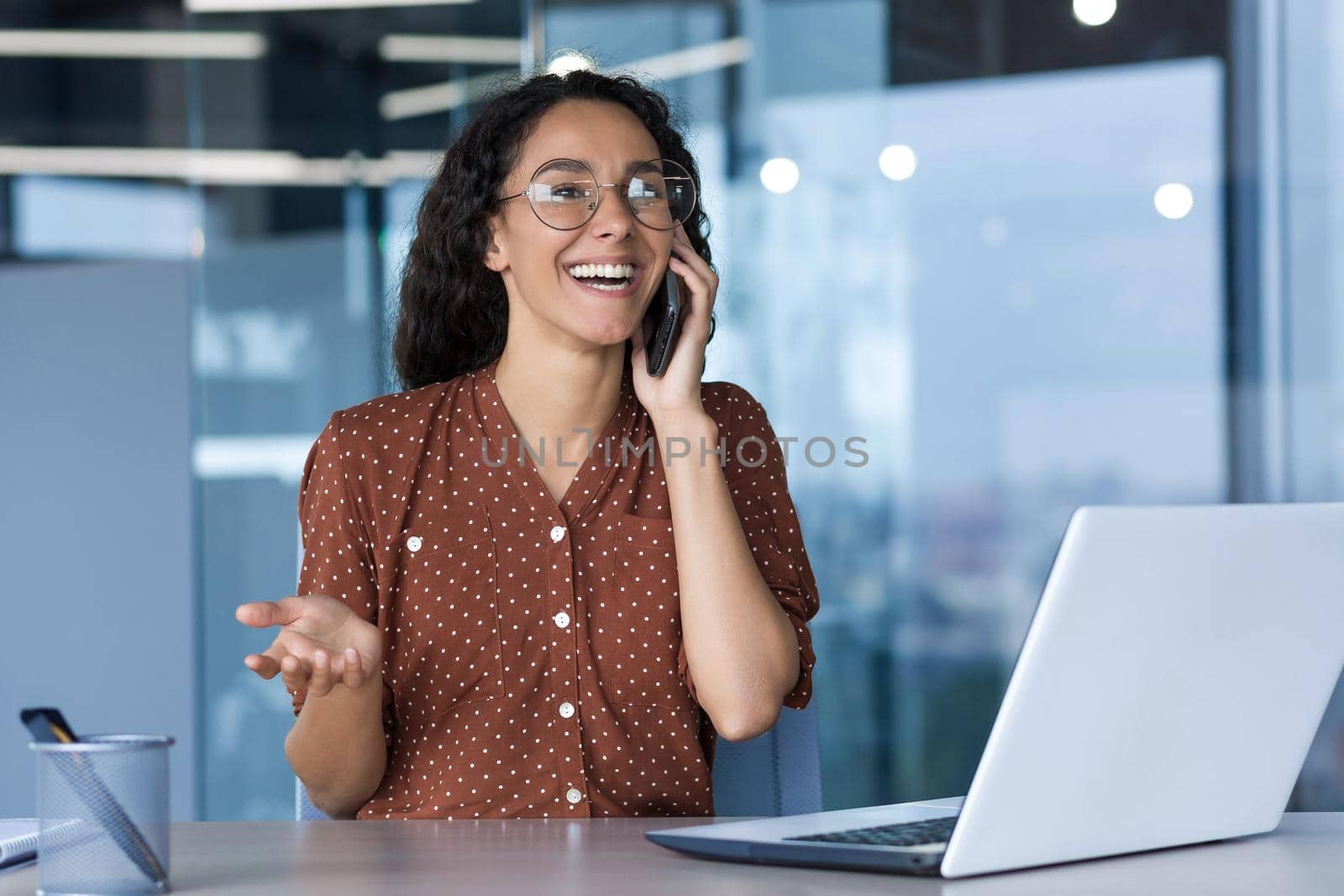 Young successful hispanic businesswoman working inside modern office, female employee cheerfully talking to colleagues on phone and smiling, using laptop while sitting at desk.