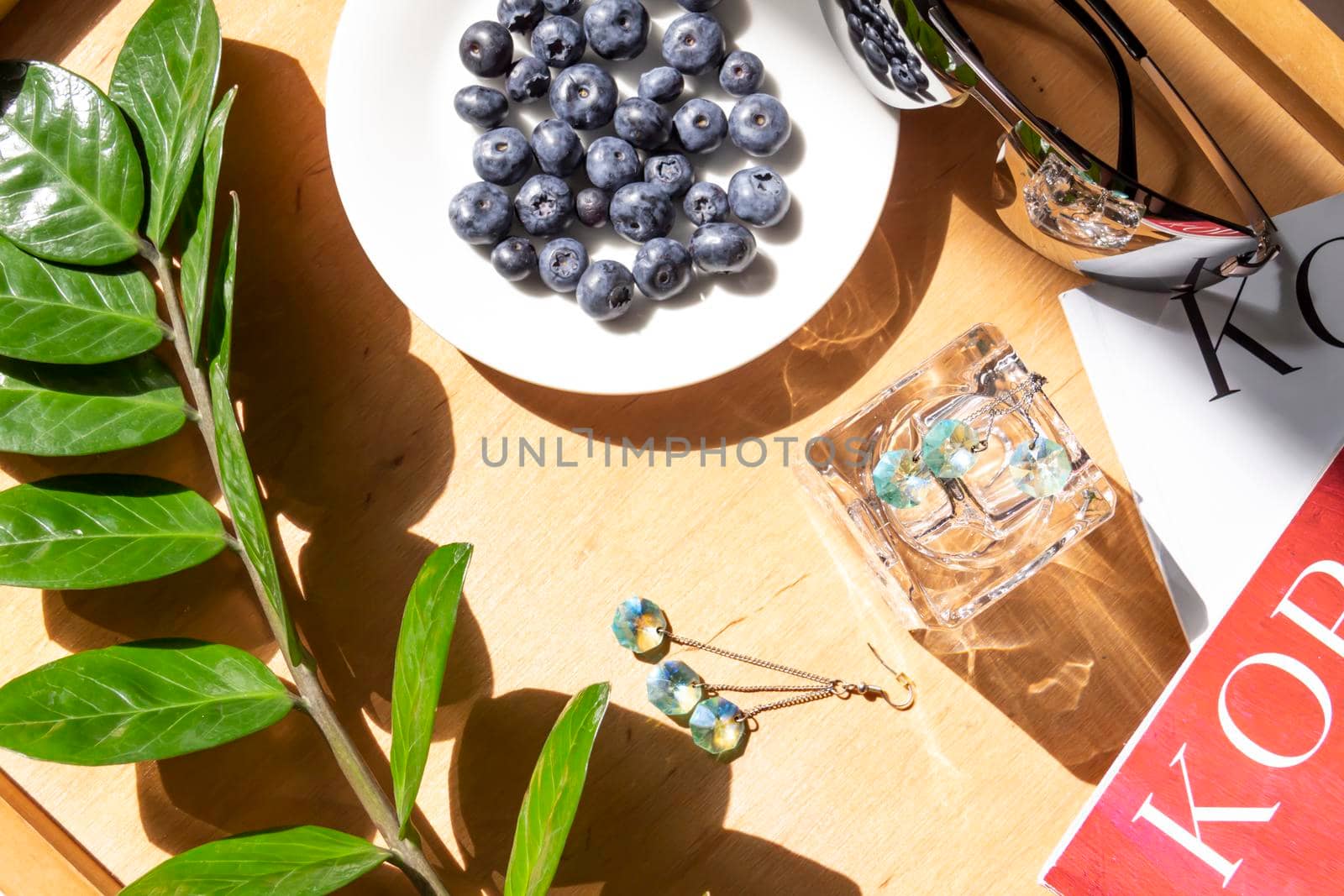 Blueberries in a white plate on a wooden table against the background of magazines and plants with green leaves.,,