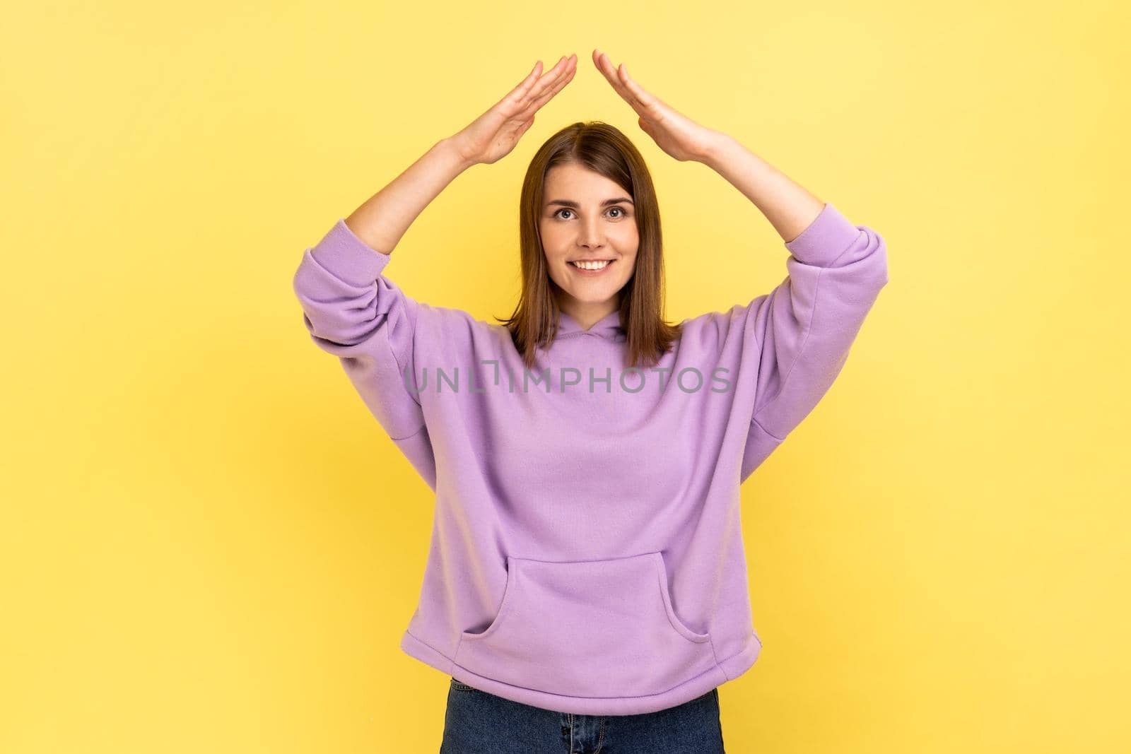 I'm in safety. Portrait of happy protected woman raising hands above head, making roof gesture, insurance, wearing purple hoodie. Indoor studio shot isolated on yellow background.