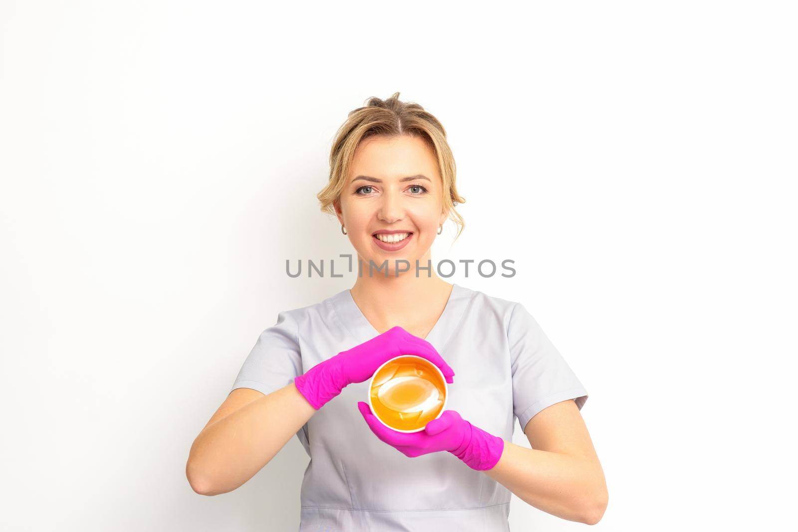 Portrait of a female caucasian beautician holding a jar of sugar paste for sugaring wearing pink gloves on white background