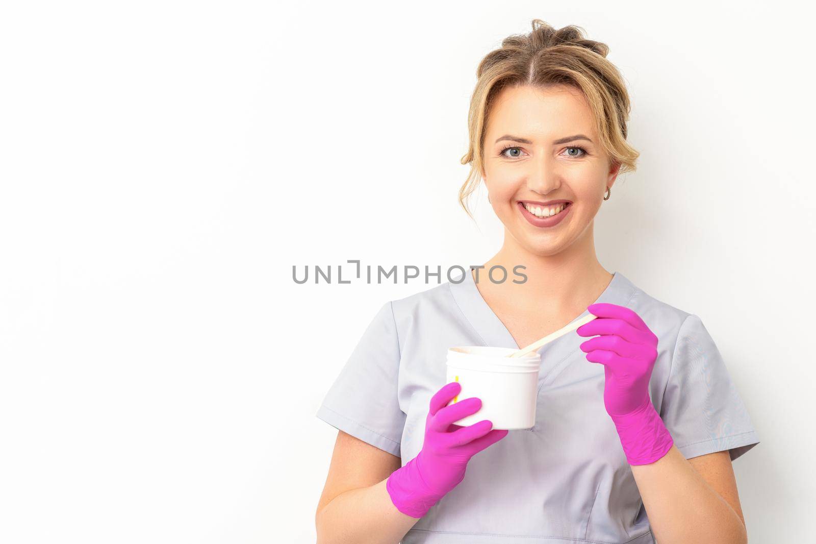 Portrait of a female caucasian beautician holding a jar of sugar paste for sugaring wearing pink gloves on white background