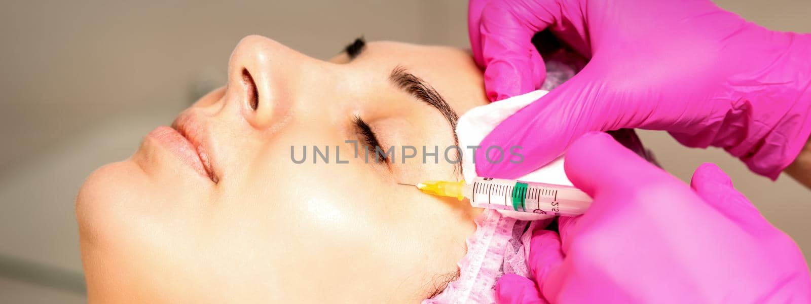 The young white woman is getting rejuvenating facial injections with hyaluronic acid on the eye in a beauty clinic