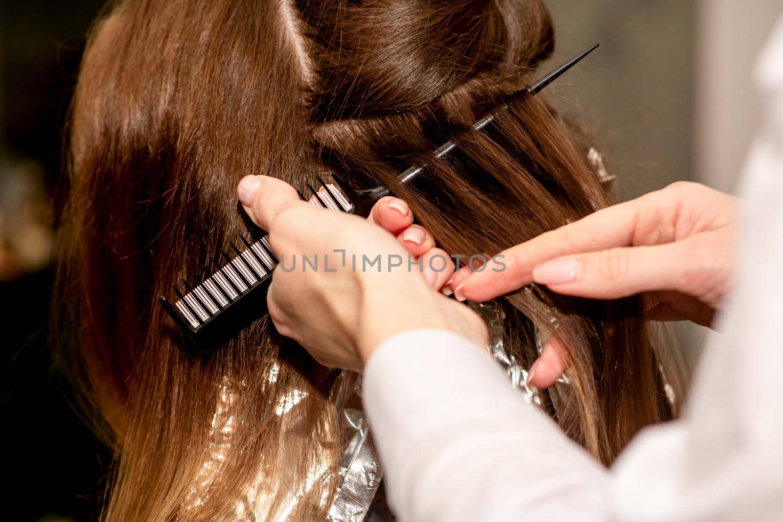 Hairdresser's hands prepare brown hair for dyeing with a comb and foil in a beauty salon, close up. by okskukuruza