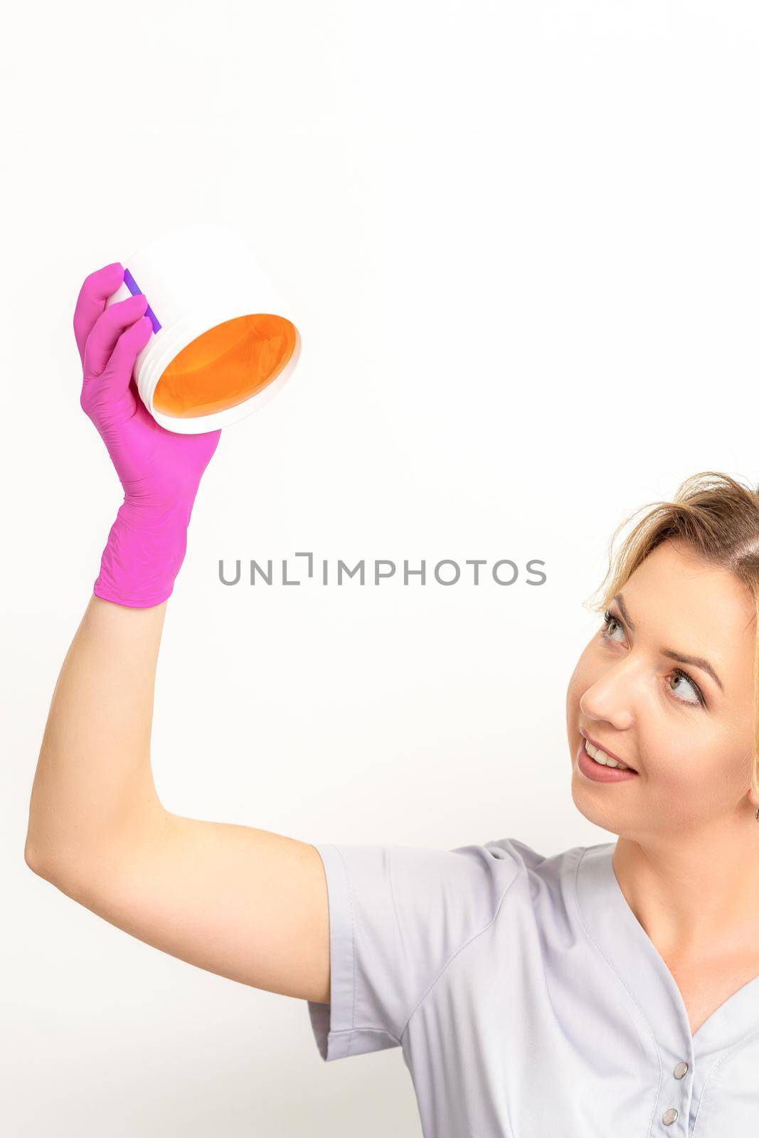 Portrait of a female caucasian beautician holding a jar of sugar paste for sugaring wearing pink gloves on white background