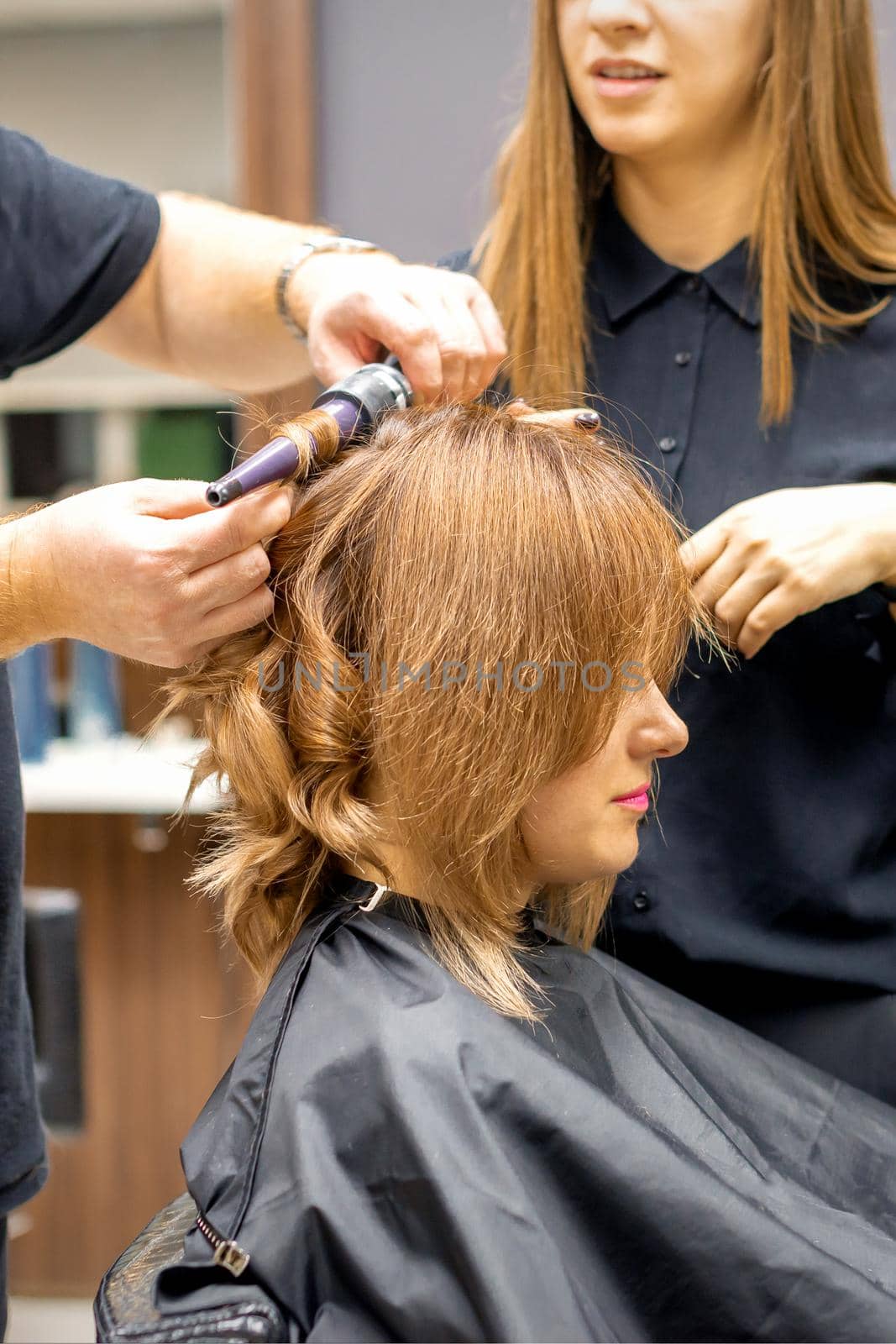 Two hairstylists make curls hairstyle of long brown hair with the curling iron in hairdresser salon. by okskukuruza