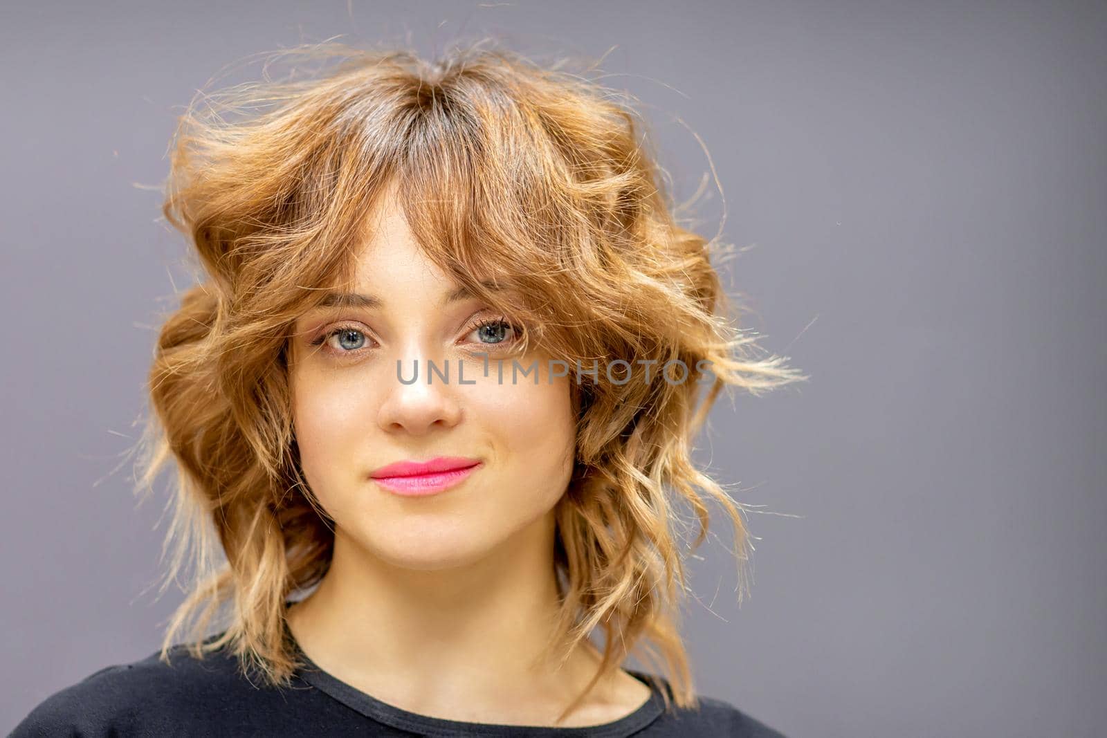 Portrait of a beautiful young caucasian red-haired woman with short wavy hairstyle smiling and looking at camera on dark gray background with copy space