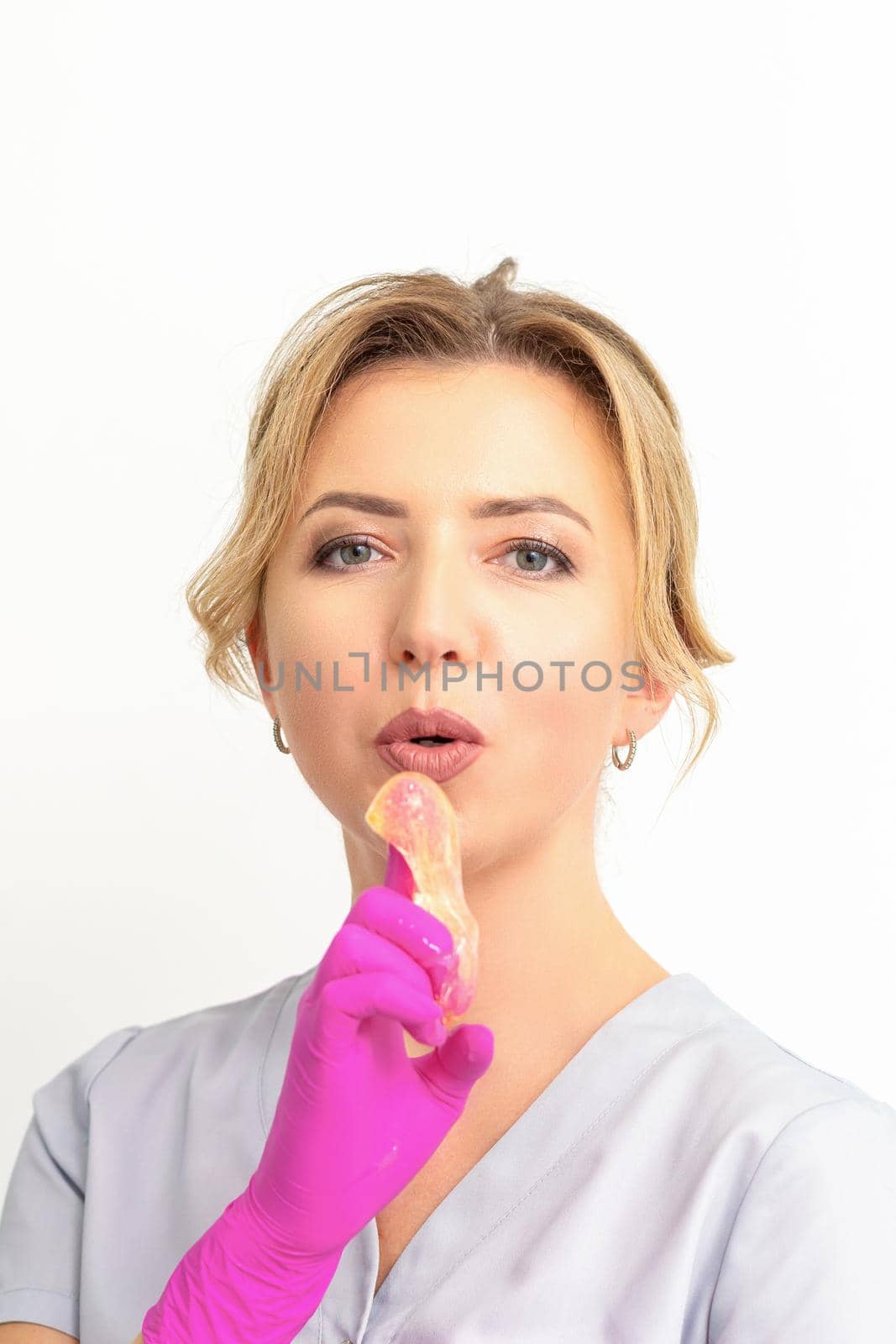 Young caucasian female beautician holding wax paste on forefinger blowing on him wearing pink gloves against a white background. Epilation concept. by okskukuruza