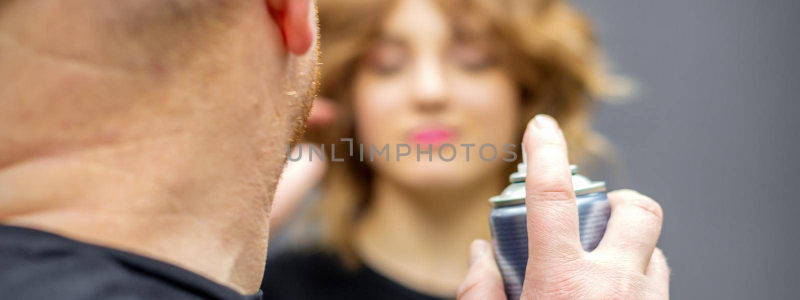 The hairdresser is using hair spray to fix the hairstyle of the young caucasian woman in the hairdresser salon, close up