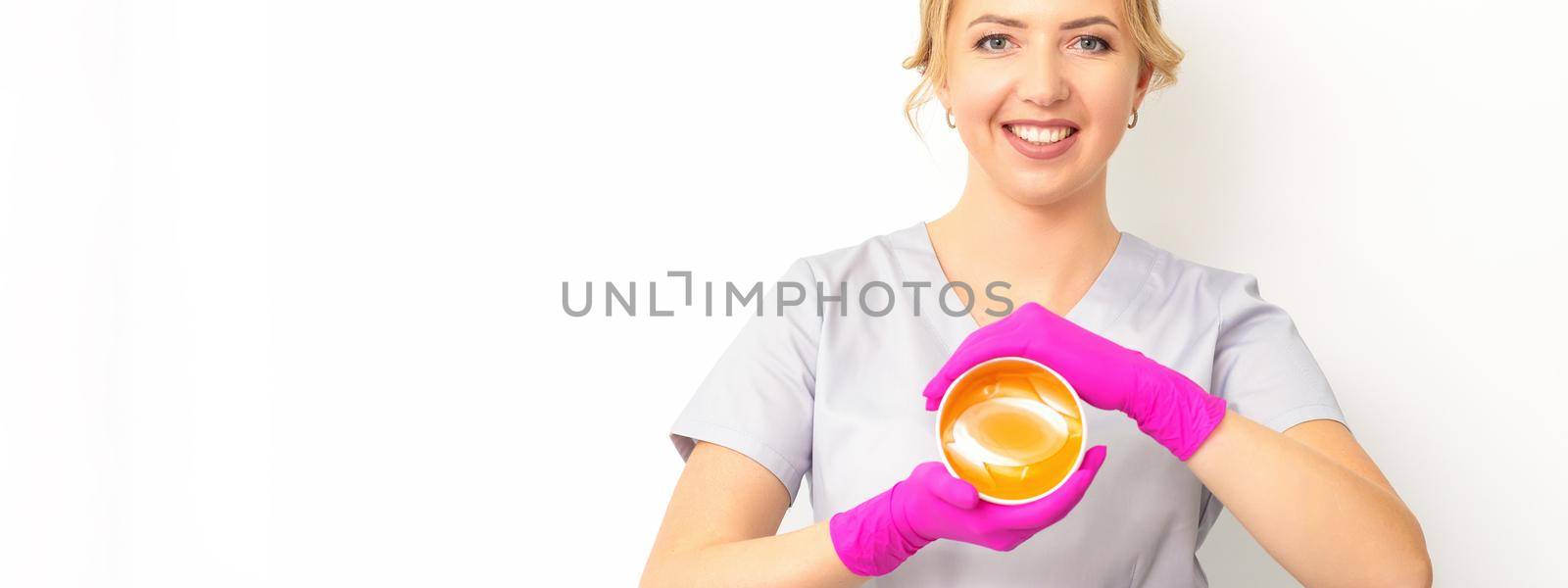 Portrait of a female caucasian beautician holding a jar of sugar paste for sugaring wearing pink gloves on white background