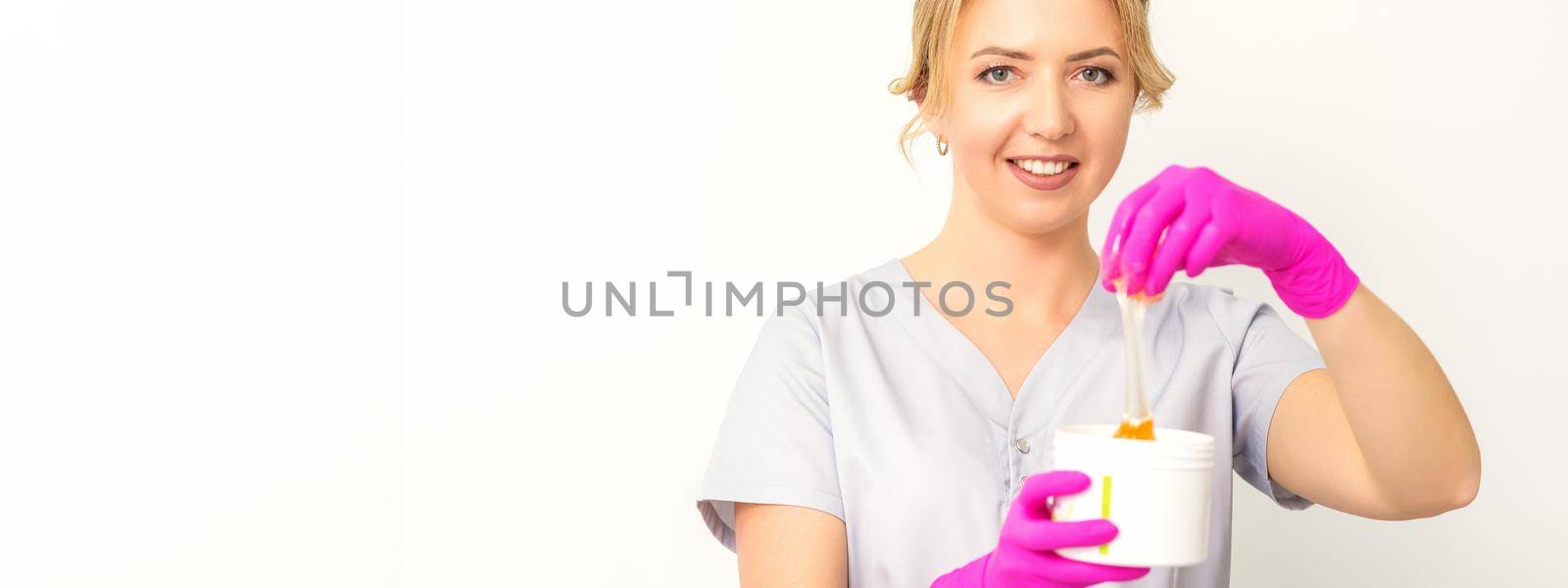 Portrait of a female caucasian beautician holding a jar of sugar paste for sugaring wearing pink gloves on white background