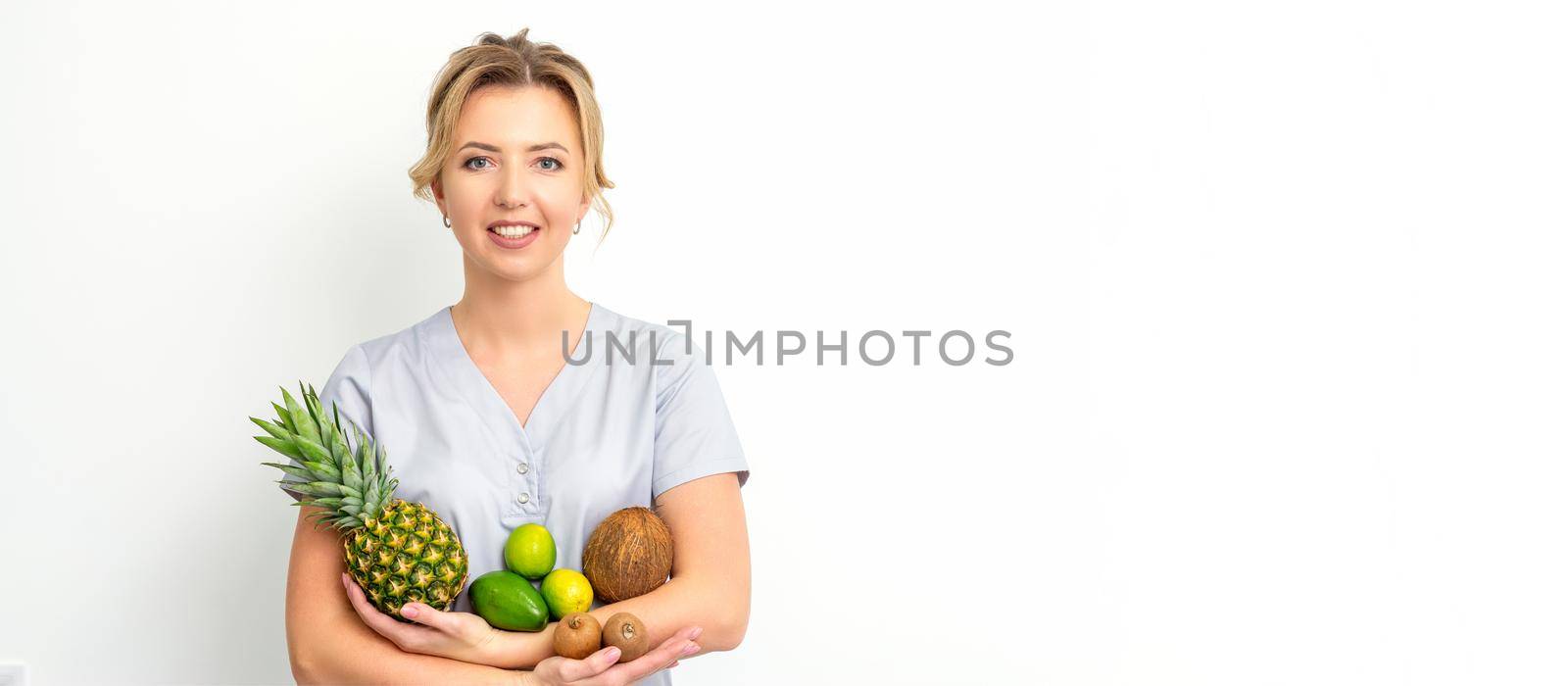Portrait of a young caucasian smiling female nutritionist with different fruits in her hands over white background. by okskukuruza