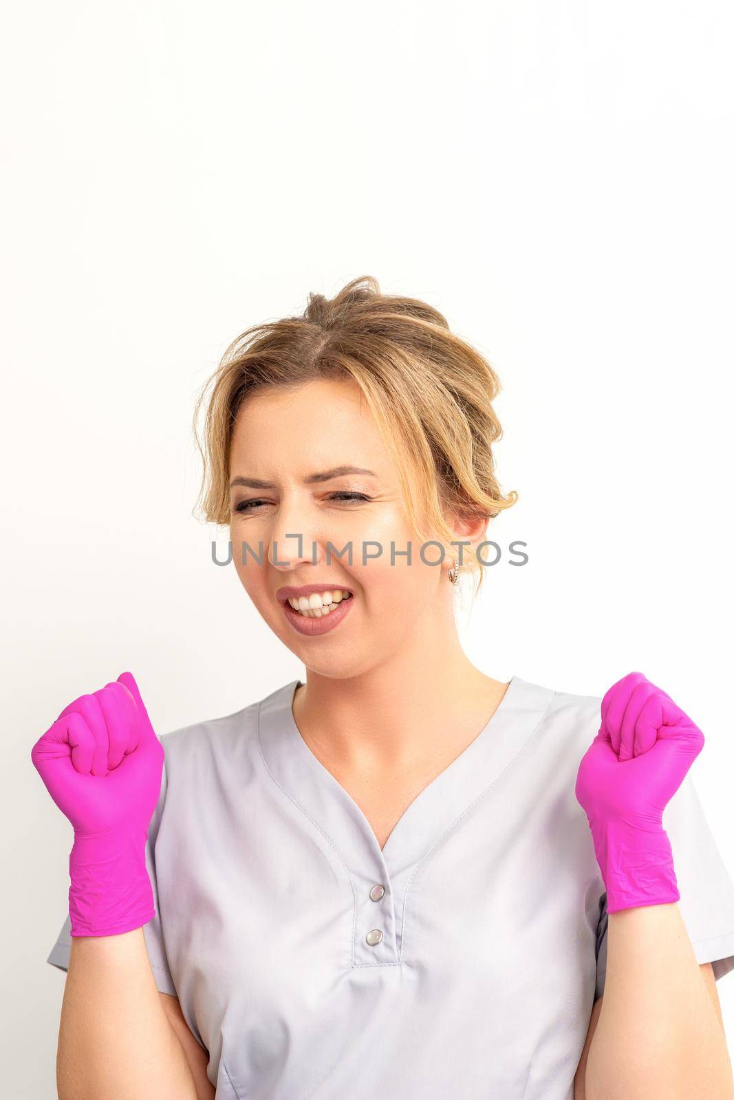 Happy caucasian woman doctor wearing pink gloves celebrates and raising fists on white background