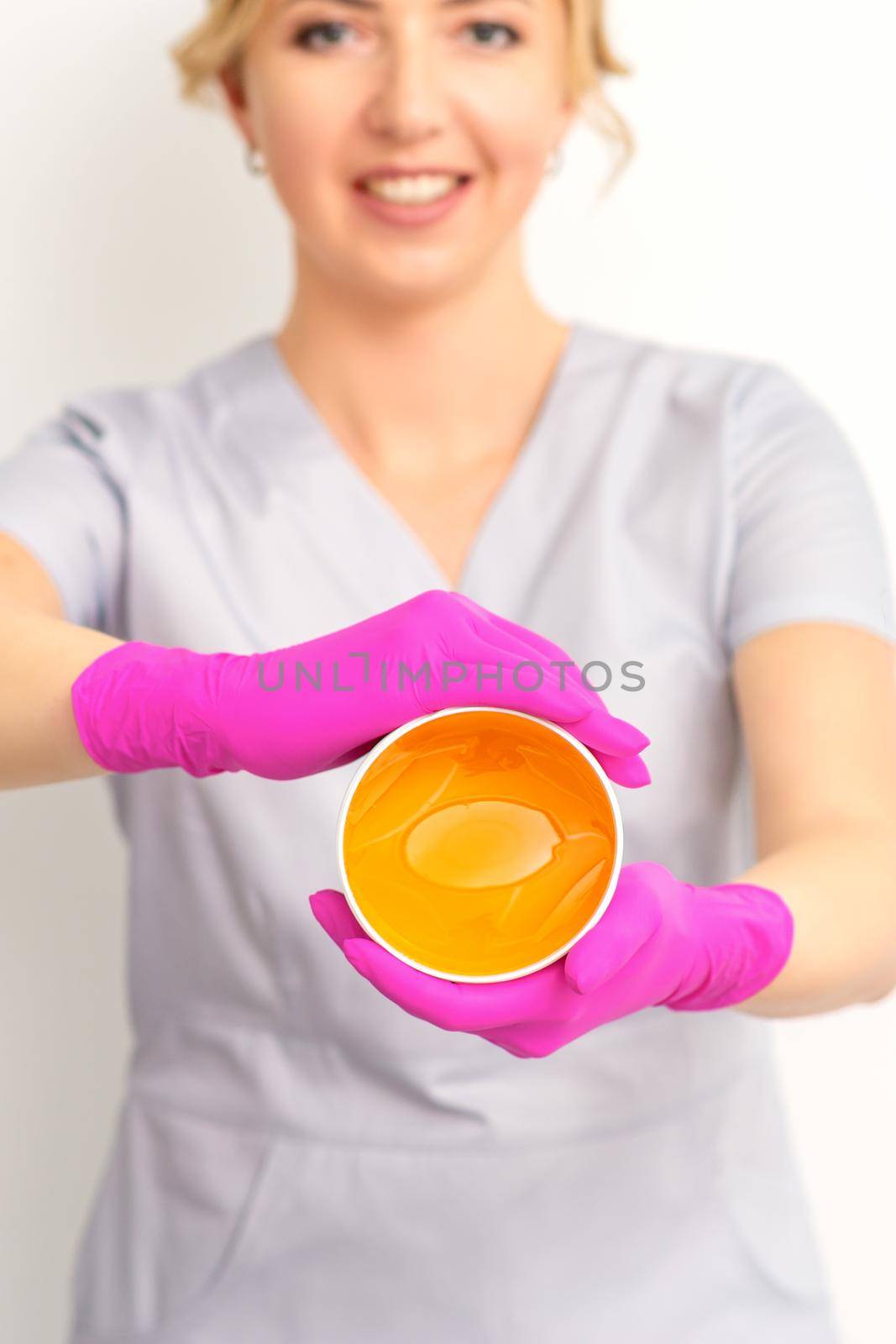 Portrait of a female caucasian beautician holding a jar of sugar paste for sugaring wearing pink gloves on white background. by okskukuruza