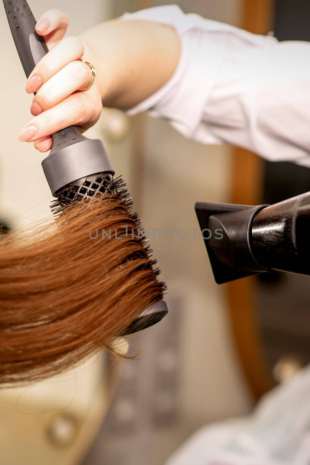 A hairdresser is drying long brown hair with a hairdryer and round brush in a beauty salon