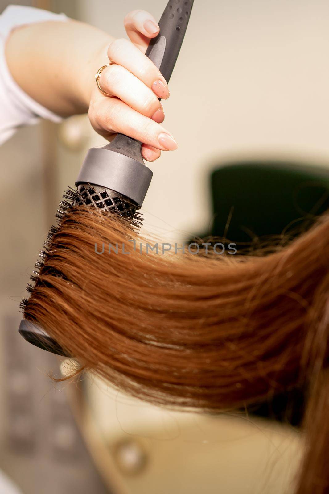 A hairdresser is drying long brown hair with a hairdryer and round brush in a beauty salon