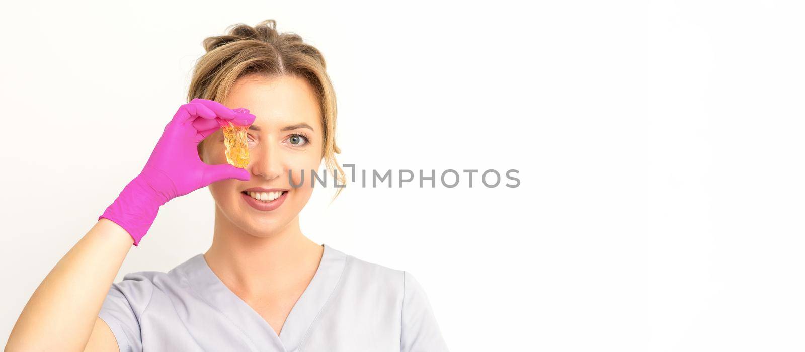 The master of sugar hair removal holds liquid yellow sugar paste, covering her eye with wax for depilation on a white background. by okskukuruza