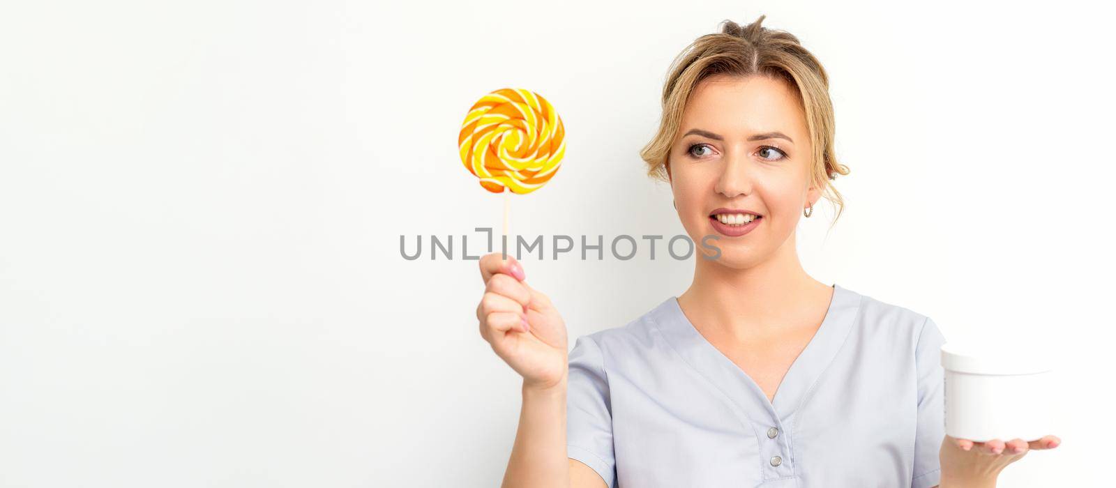 Waxing, depilation concept. The beautician is wearing a medical coat holding a sugar lollipop with a jar of sugar paste on white background. by okskukuruza