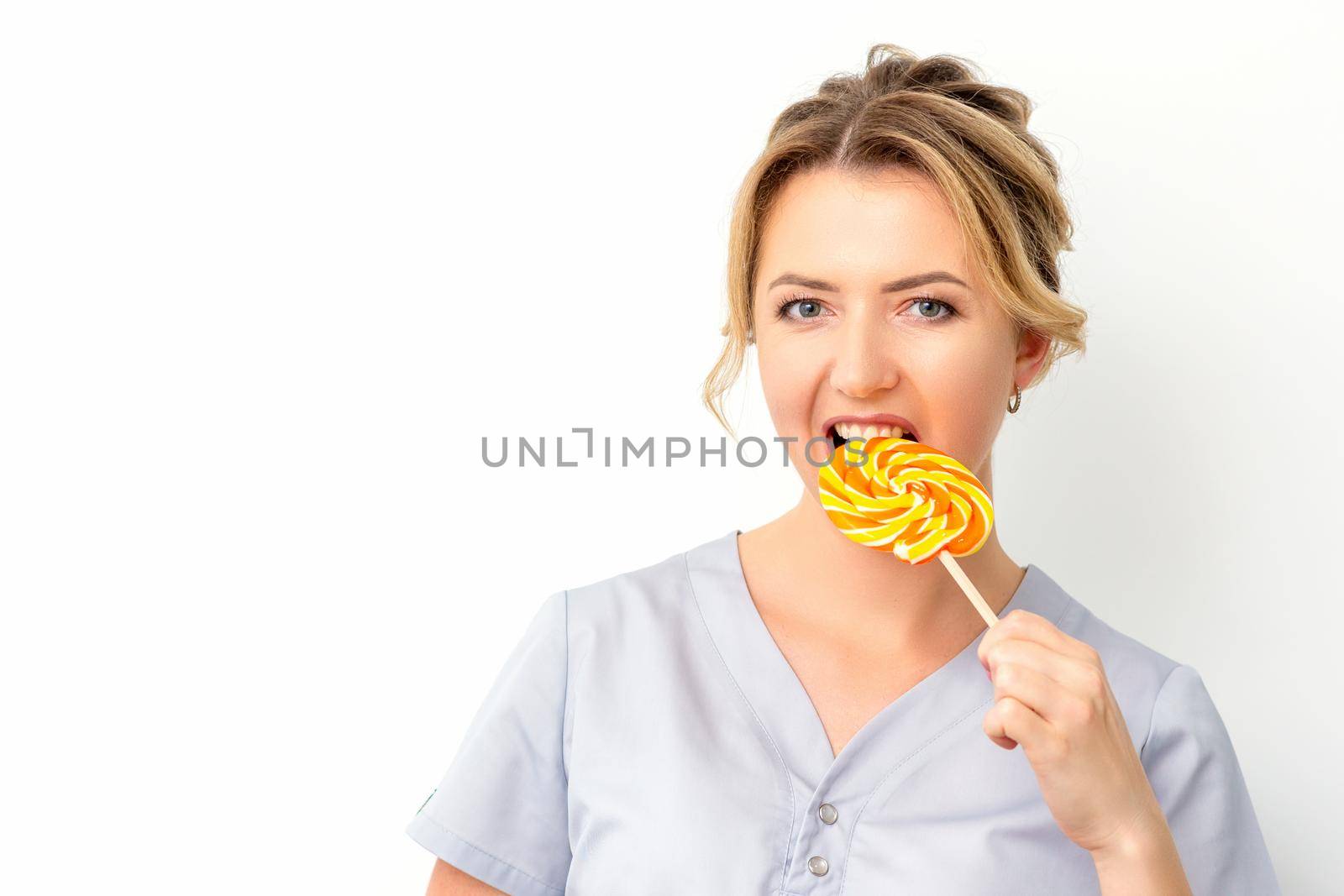 Portrait of a beautiful young caucasian beautician wearing a medical shirt bites a lollipop on a white background. by okskukuruza