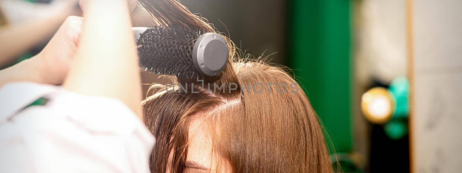 A hairdresser is drying long brown hair with a hairdryer and round brush in a beauty salon. by okskukuruza