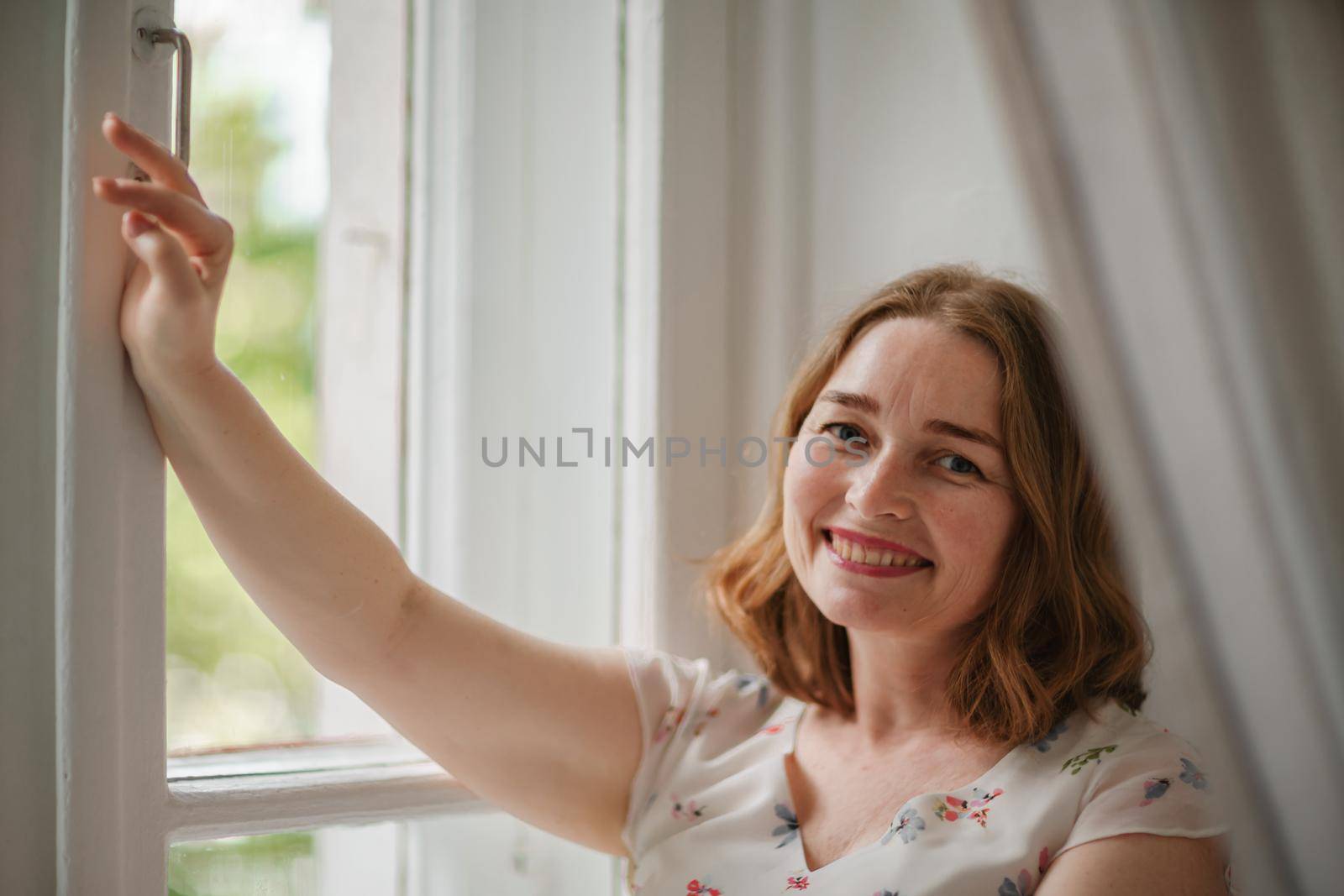 A middle-aged woman in a cream dress sits mysteriously and looks out the window on the windowsill. Green trees outside