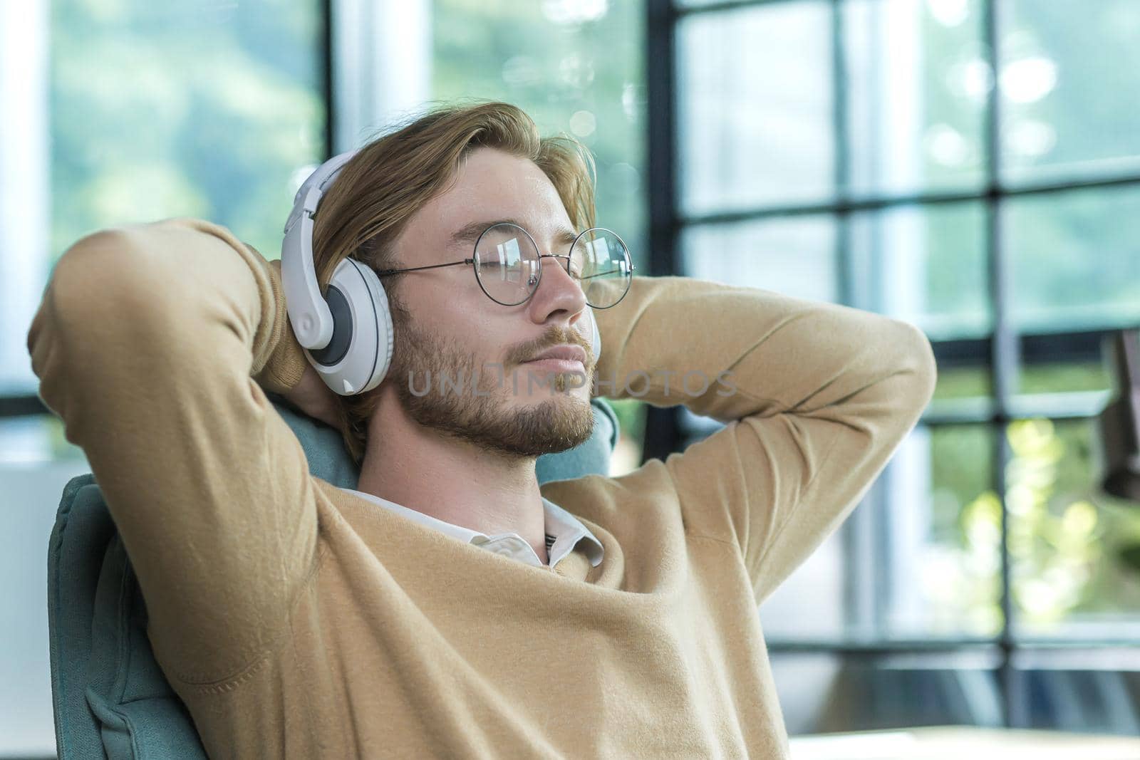 A man in an office on a chair with his arms folded is listening to music in headphones resting, a blond businessman is working inside an office building